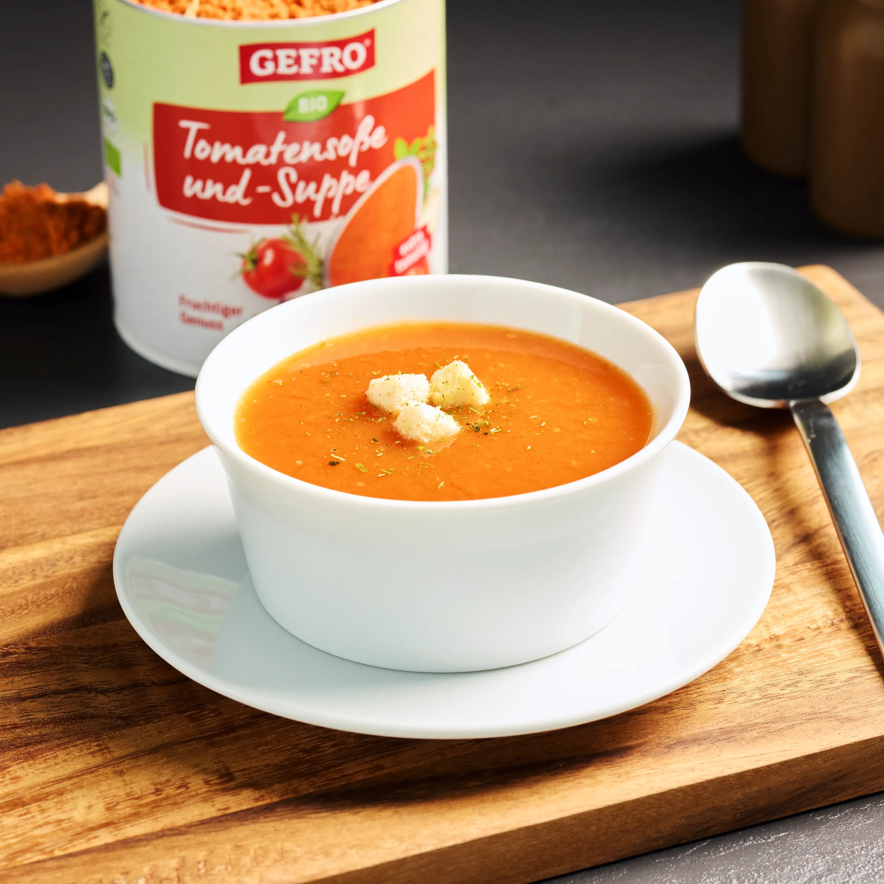 Schüssel mit cremiger Tomatensuppe auf Untertasse, garniert mit Croutons; dahinter Dose von Gefro Bio Tomatensuppe