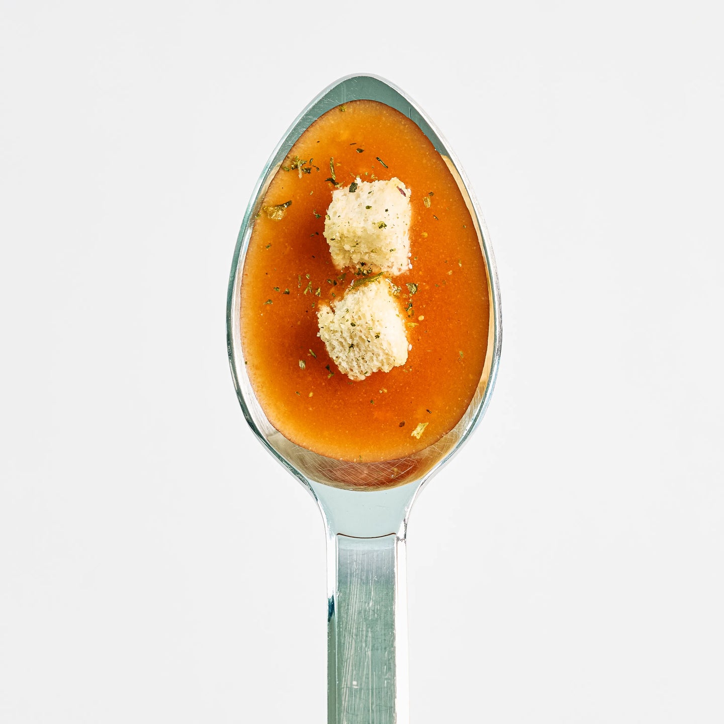 Löffel mit cremiger roter Tomatensuppe, garniert mit zwei Brotwürfeln und Kräutern, vor hellem Hintergrund