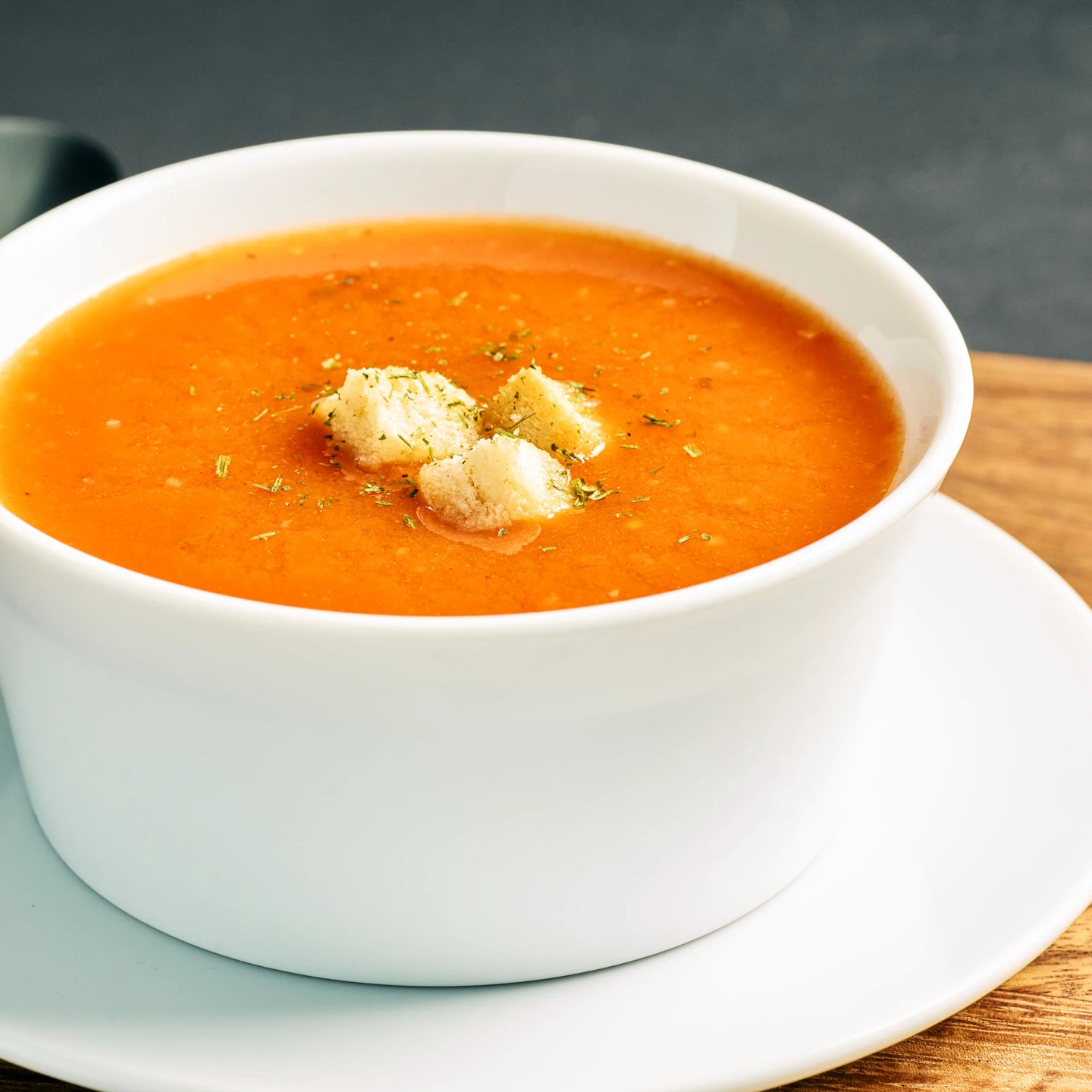 Weiße Schale mit cremiger Tomatensuppe, garniert mit Croutons und Kräutern, serviert auf Untertasse.