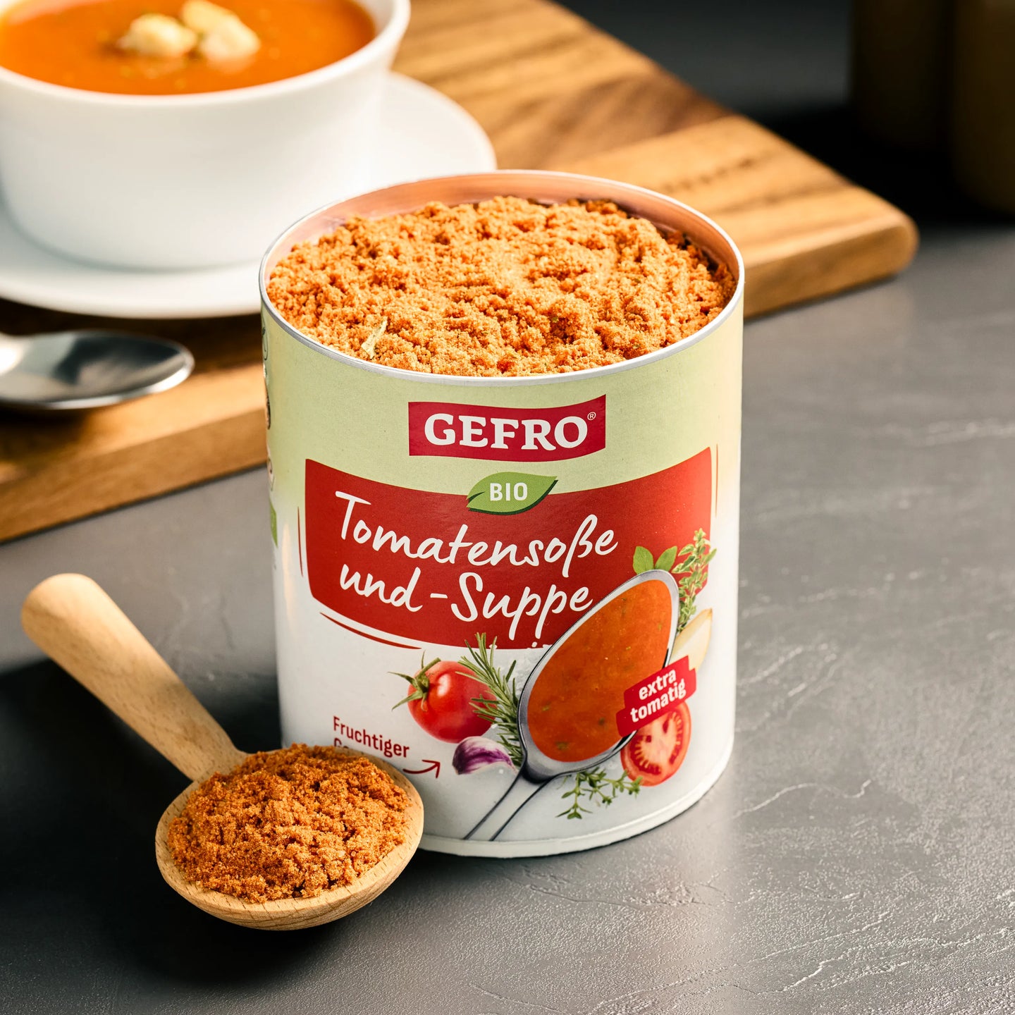 Offene Dose GEFRO Bio Tomatensoße und -Suppe mit Pulver im Holzlöffel, daneben eine servierte Tomatensuppe.