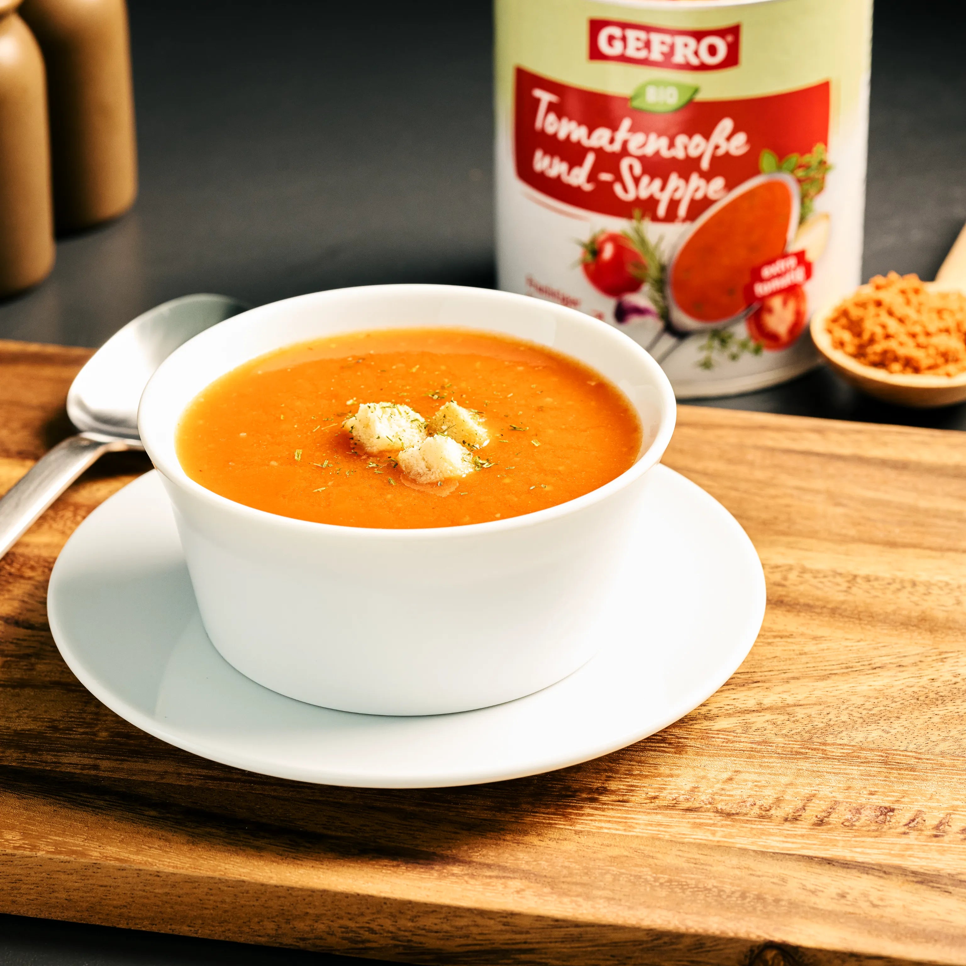 Weiße Schale mit Tomatensuppe und Croûtons auf Holzbrett, dahinter Dose Gefro Bio Tomatensoße und -Suppe.