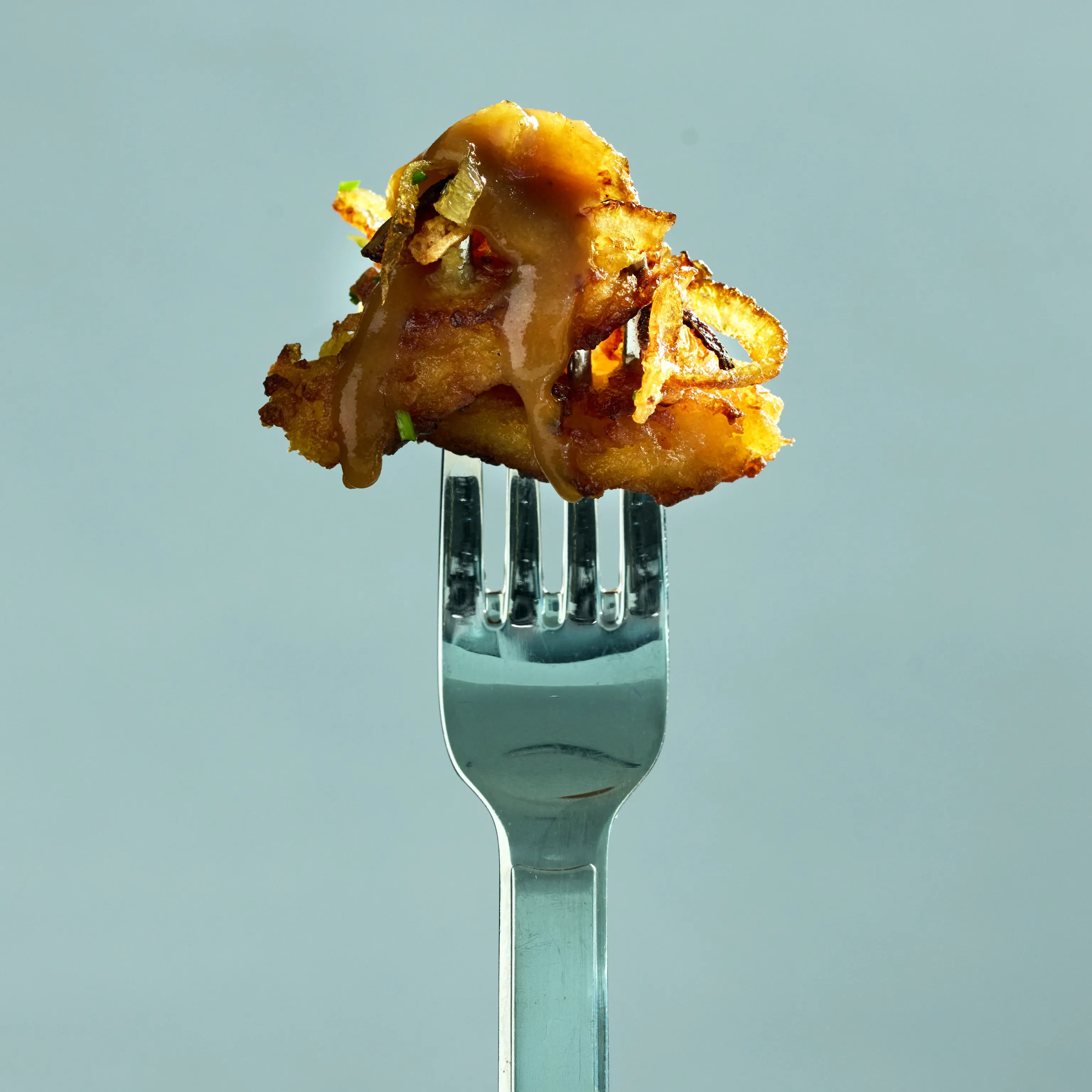 Knuspriger, goldbrauner Happen auf einer Gabel, mit brauner Sauce und gerösteten Zwiebeln vor hellem Hintergrund