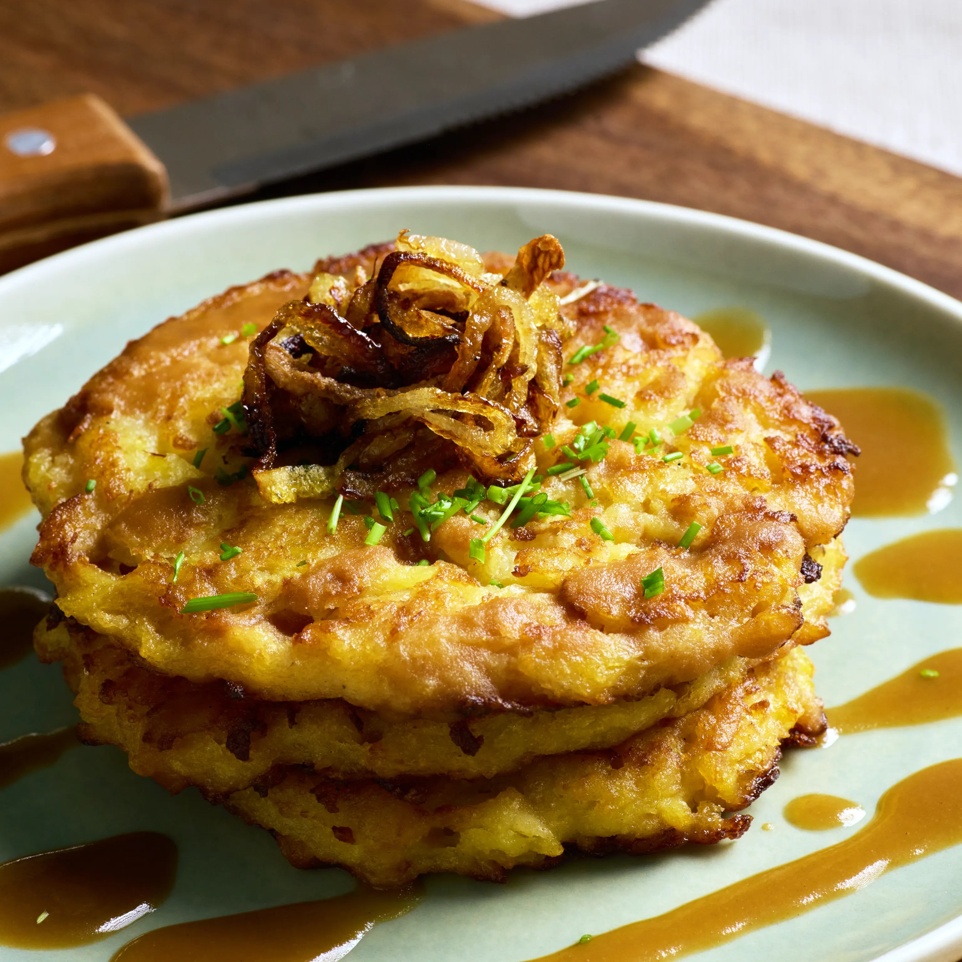Drei knusprige Kartoffelpuffer auf einem Teller, garniert mit Röstzwiebeln, Schnittlauch und brauner Sauce.