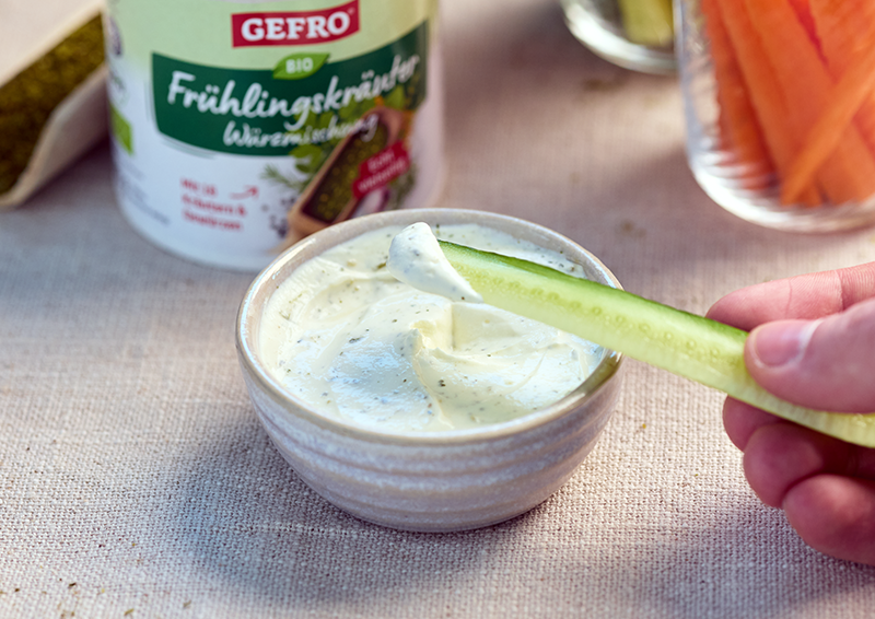 Gurkenstift wird in cremigen Kräuter-Dip getaucht, daneben steht ein Becher GEFRO Bio Frühlingskräuter Würzmischung.