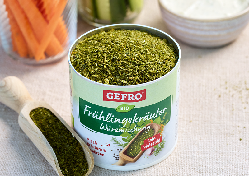 Dose GEFRO Bio Frühlingskräuter Würzmischung mit geöffnetem Inhalt und Holzschaufel auf heller Unterlage