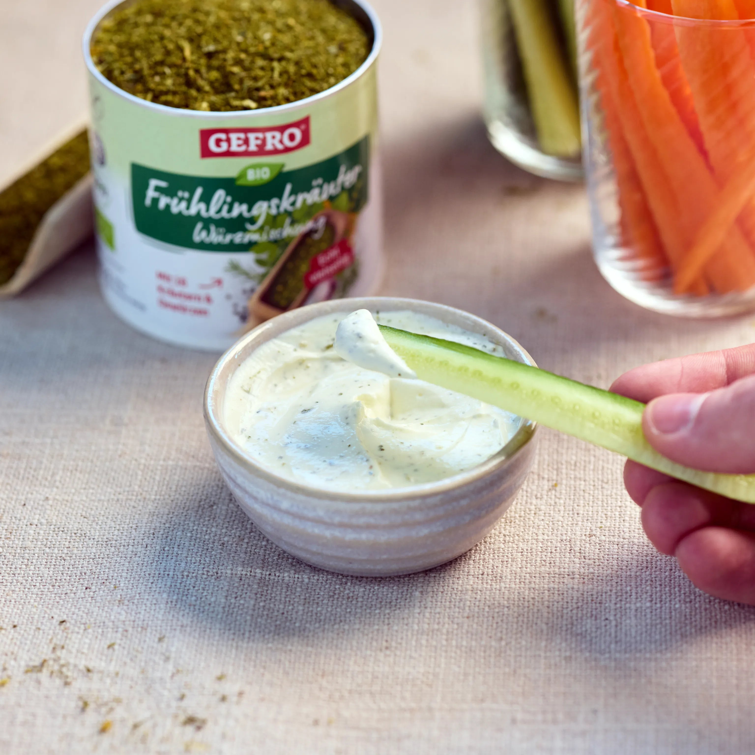 Gurkenscheibe wird in einen cremigen Kräuterdip getaucht, daneben eine Dose GEFRO Bio Frühlingskräuter.
