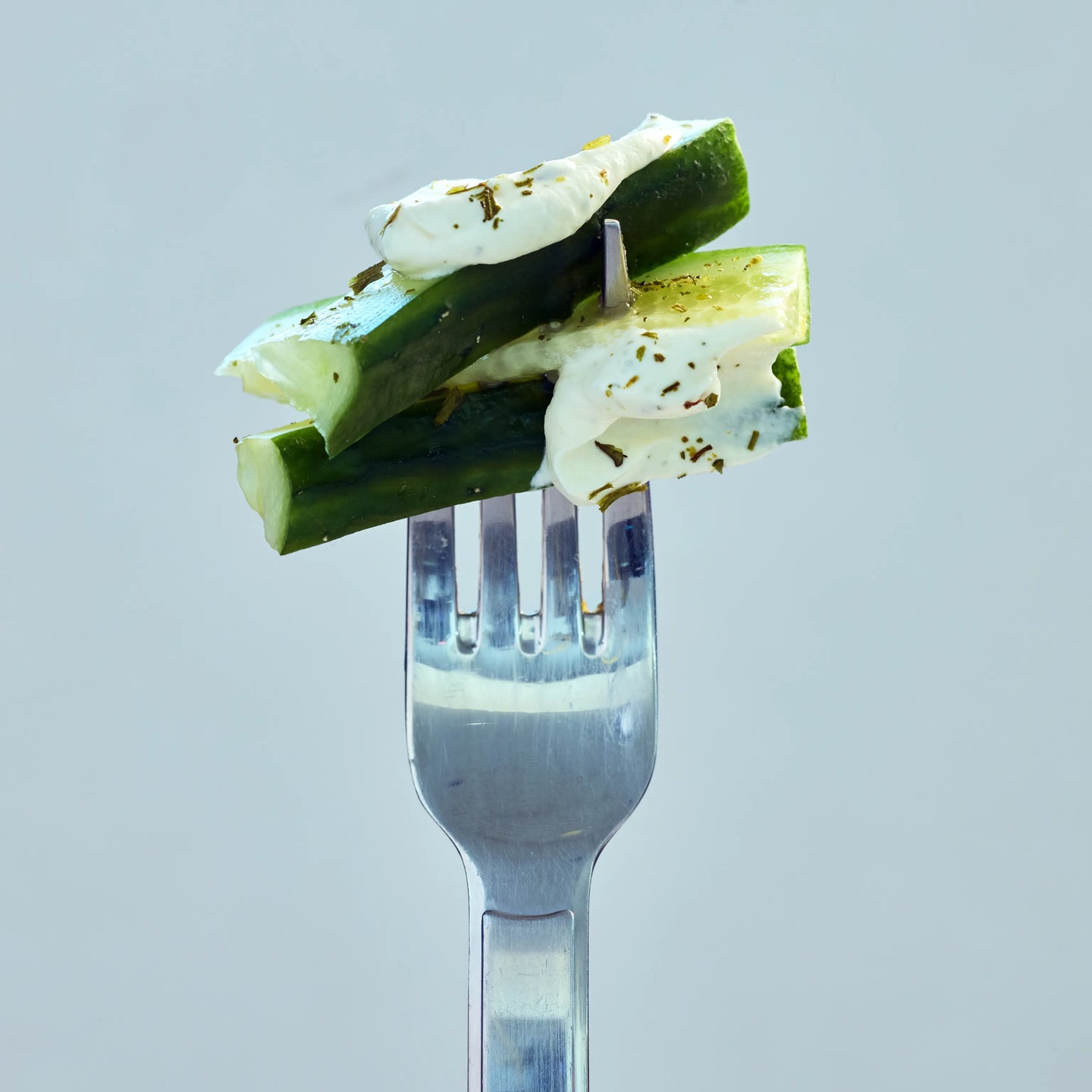 Gurkenstücke mit cremigem Dip und Kräutern auf einer Gabel, als frischer Snack vor hellem Hintergrund präsentiert