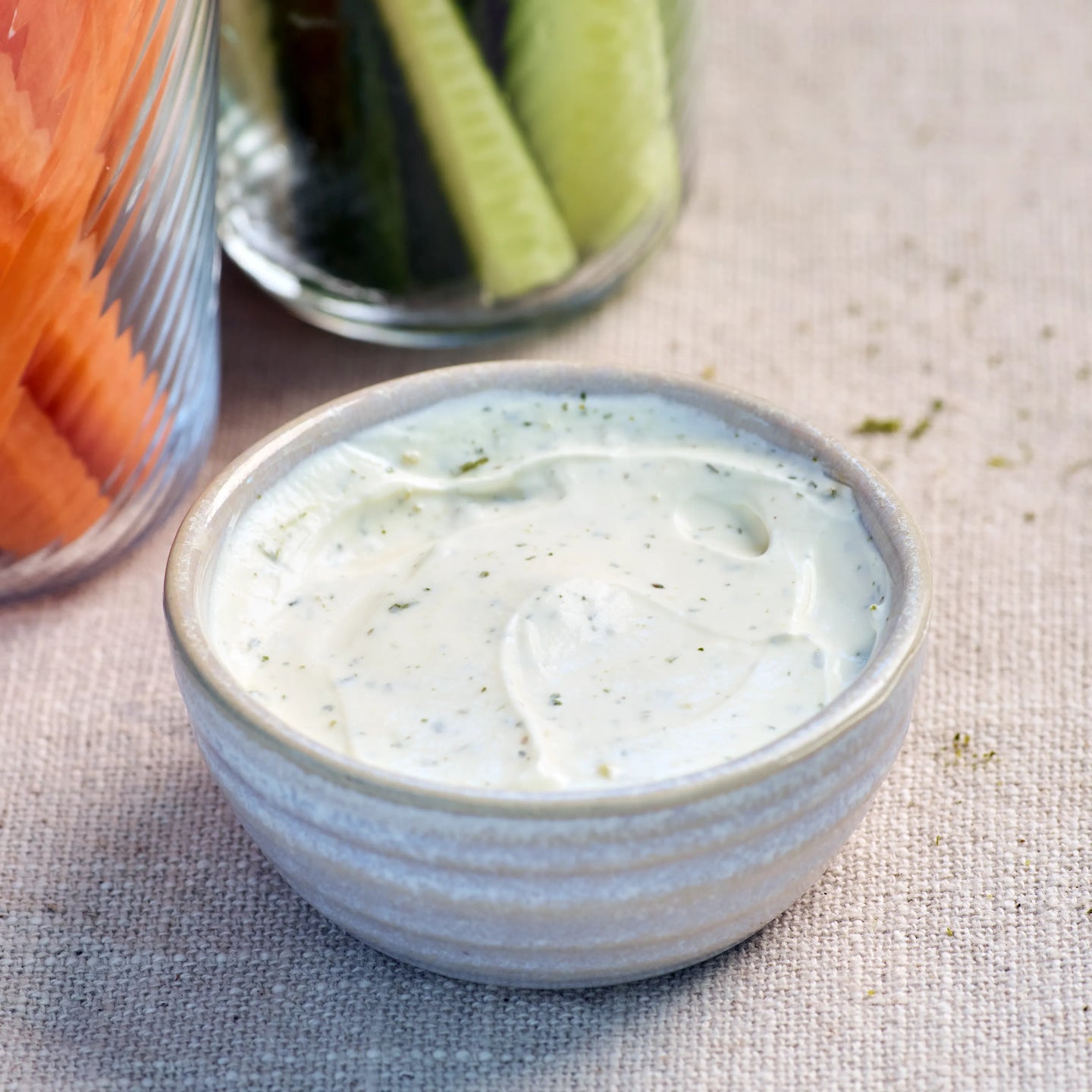 Kleine Schale mit cremigem Kräuterdip auf hellem Stoff, daneben Gemüsesticks im Glas als Serviervorschlag