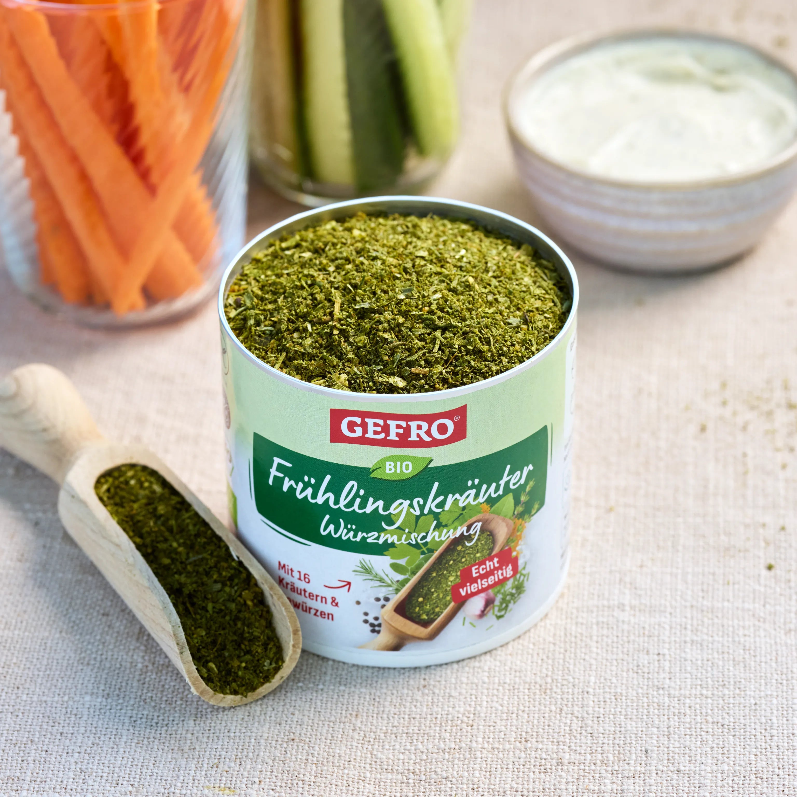 Offene Dose mit grüner Frühlingskräuter-Würzmischung von Gefro, daneben Holzschaufel und Gemüsesticks mit Dip