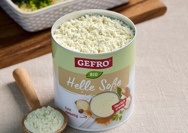 Dose GEFRO Bio Helle Soße mit cremigem Pulver, daneben ein Holzlöffel mit Soßenpulver auf hellem Tisch