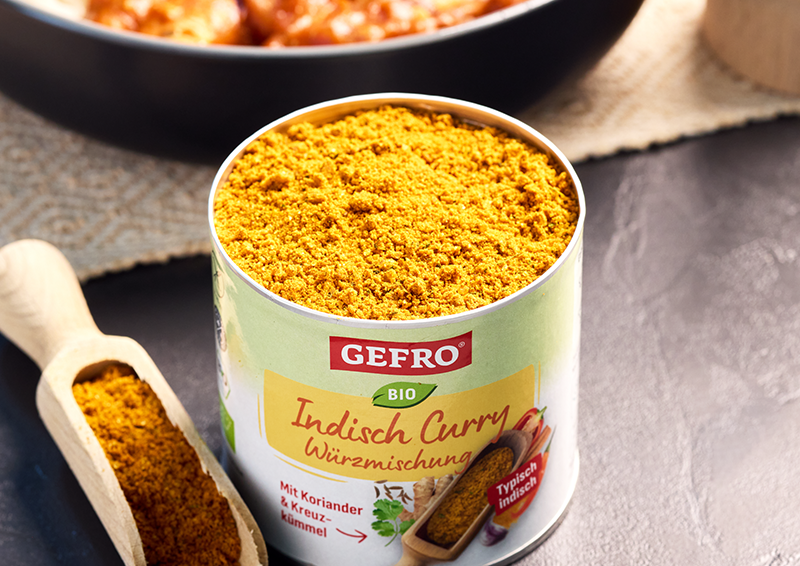 Dose mit GEFRO Bio Indisch Curry Würzmischung, geöffnet und gefüllt, daneben Holzschaufel mit Gewürzpulver