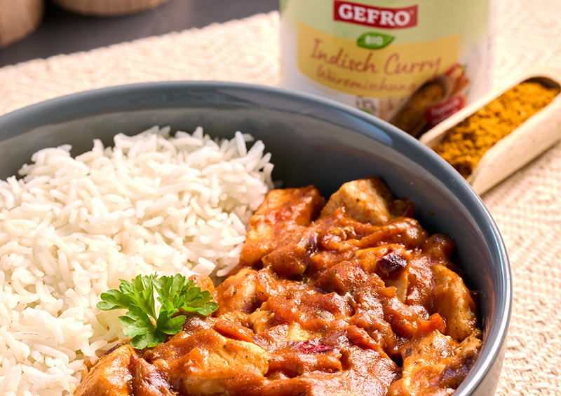 Schale mit weißem Reis und cremigem Curry mit Hähnchen, dekoriert mit Petersilie, daneben Currypulver und Dose