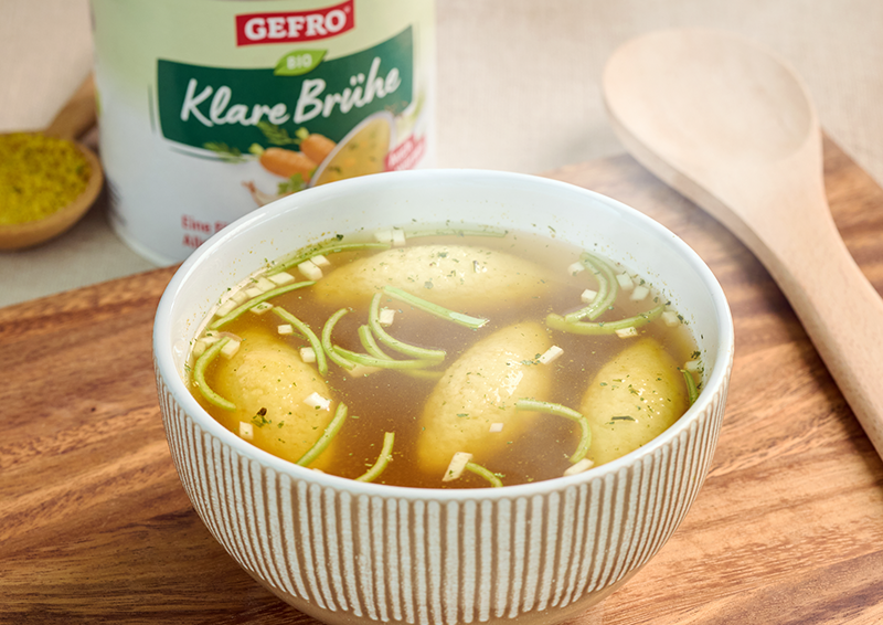 Schüssel mit klarer Brühe und vier Grießklößchen auf Holzbrett, dahinter Gefro Bio Klare Brühe und Kochlöffel