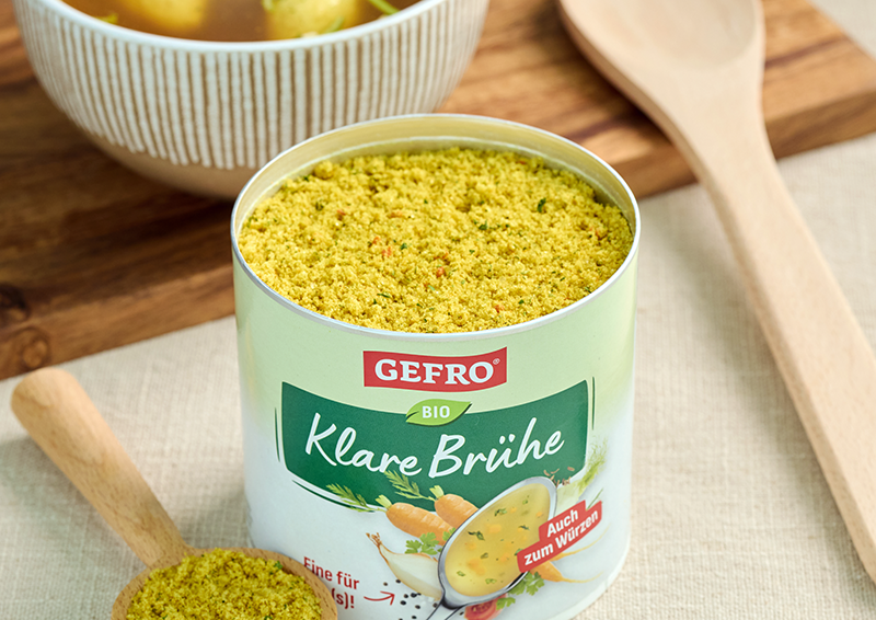 Offene Dose GEFRÖ Bio Klare Brühe mit gelbem Pulver auf Tisch, daneben Holzlöffel für Küchengebrauch