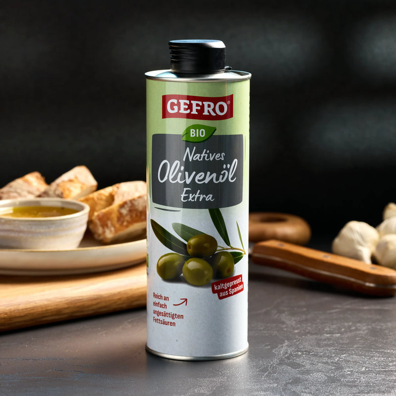 Dose GEFRÖ Bio natives Olivenöl extra auf Tisch, daneben Brot, Schale mit Öl und Knoblauch im Hintergrund