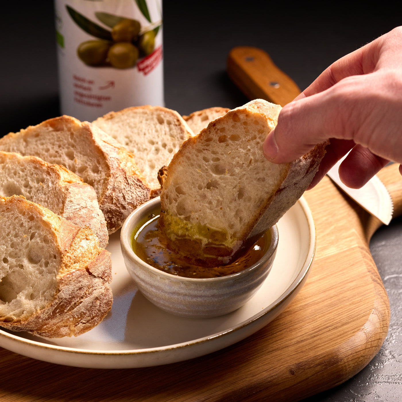 Hand taucht eine Scheibe Weißbrot in Olivenöl mit Gewürzen, daneben weitere Brotscheiben auf einem Teller.