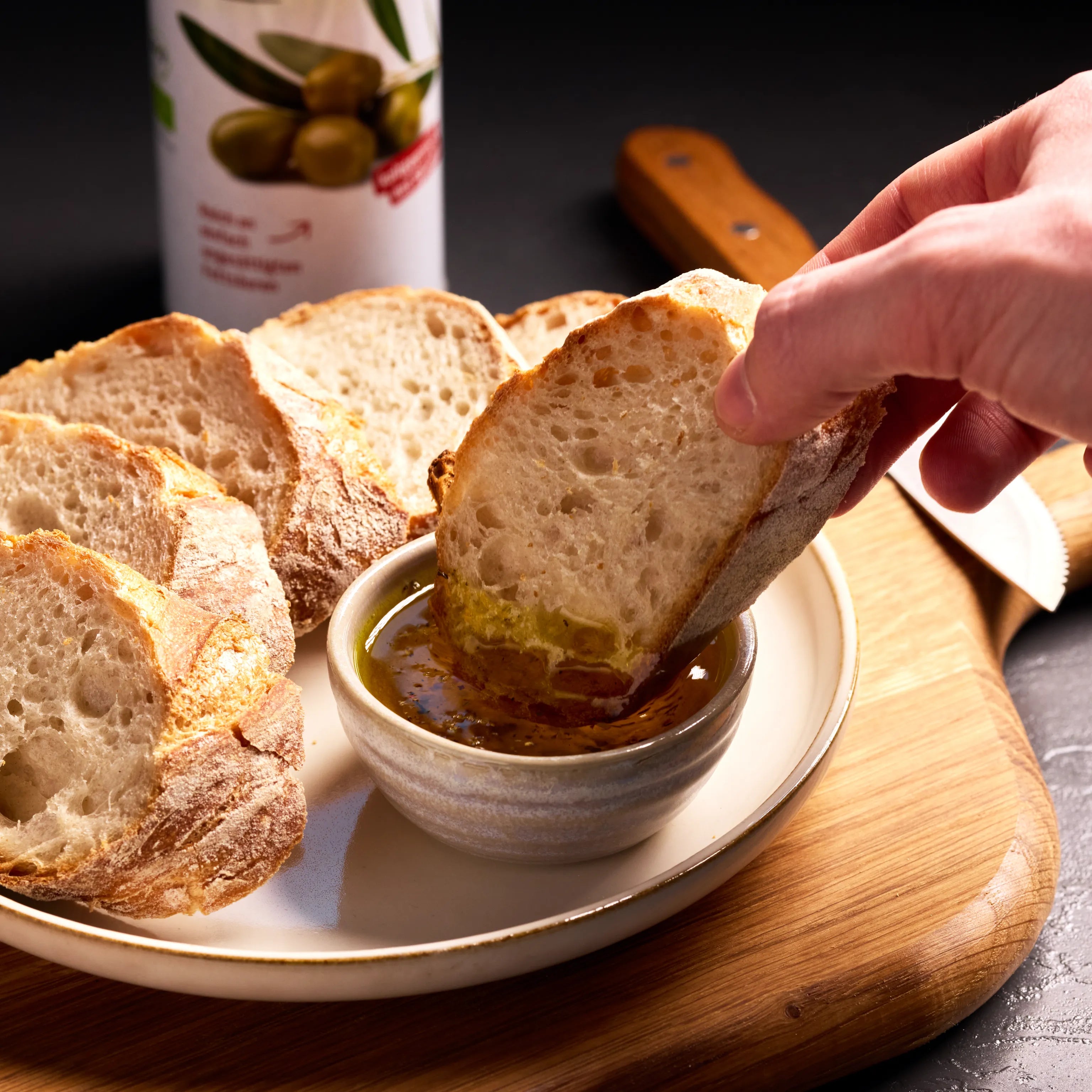 Hand taucht eine Scheibe rustikales Brot in Olivenöl mit Kräutern, serviert auf Teller mit Brotauswahl.