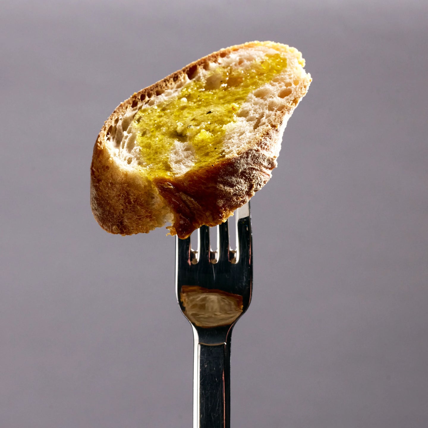 Nahaufnahme einer Brotscheibe mit goldgelbem Olivenöl auf einer Gabel vor schlichtem grauem Hintergrund