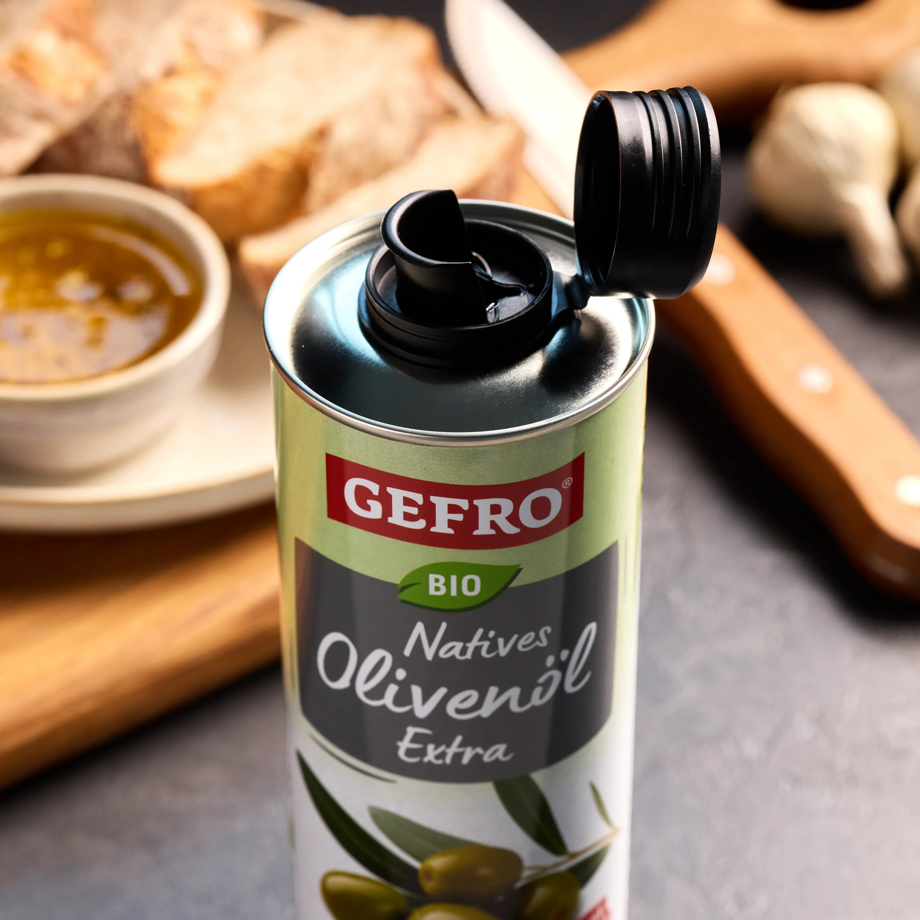 Geöffnete Dose GEFRO Bio natives Olivenöl extra auf Küchentisch, daneben Brot, Knoblauch und eine Schale Suppe