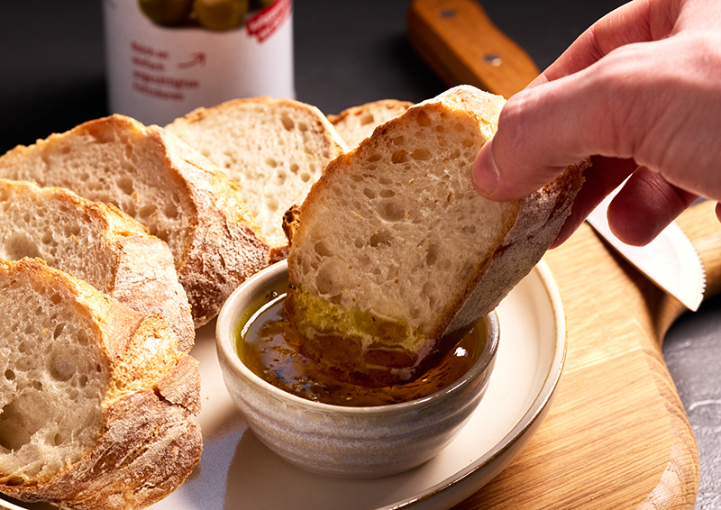 Hand taucht eine Scheibe Bauernbrot in Olivenöl mit Gewürzen, serviert auf Teller mit weiterem aufgeschnittenem Brot.