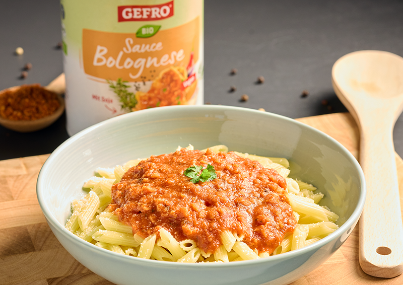 Schüssel mit Penne und Bolognese-Sauce auf Holzbrett, dahinter GEFRO Bio Sauce Bolognese und Kochlöffel