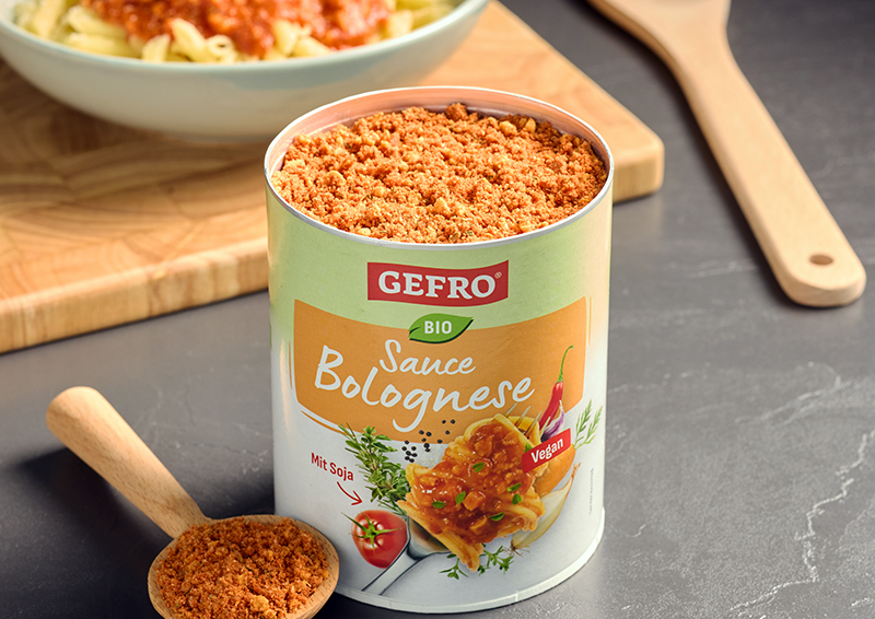 Dose GEFRO Bio Sauce Bolognese mit veganem Pulver, daneben Holzlöffel und im Hintergrund ein Teller mit Pasta
