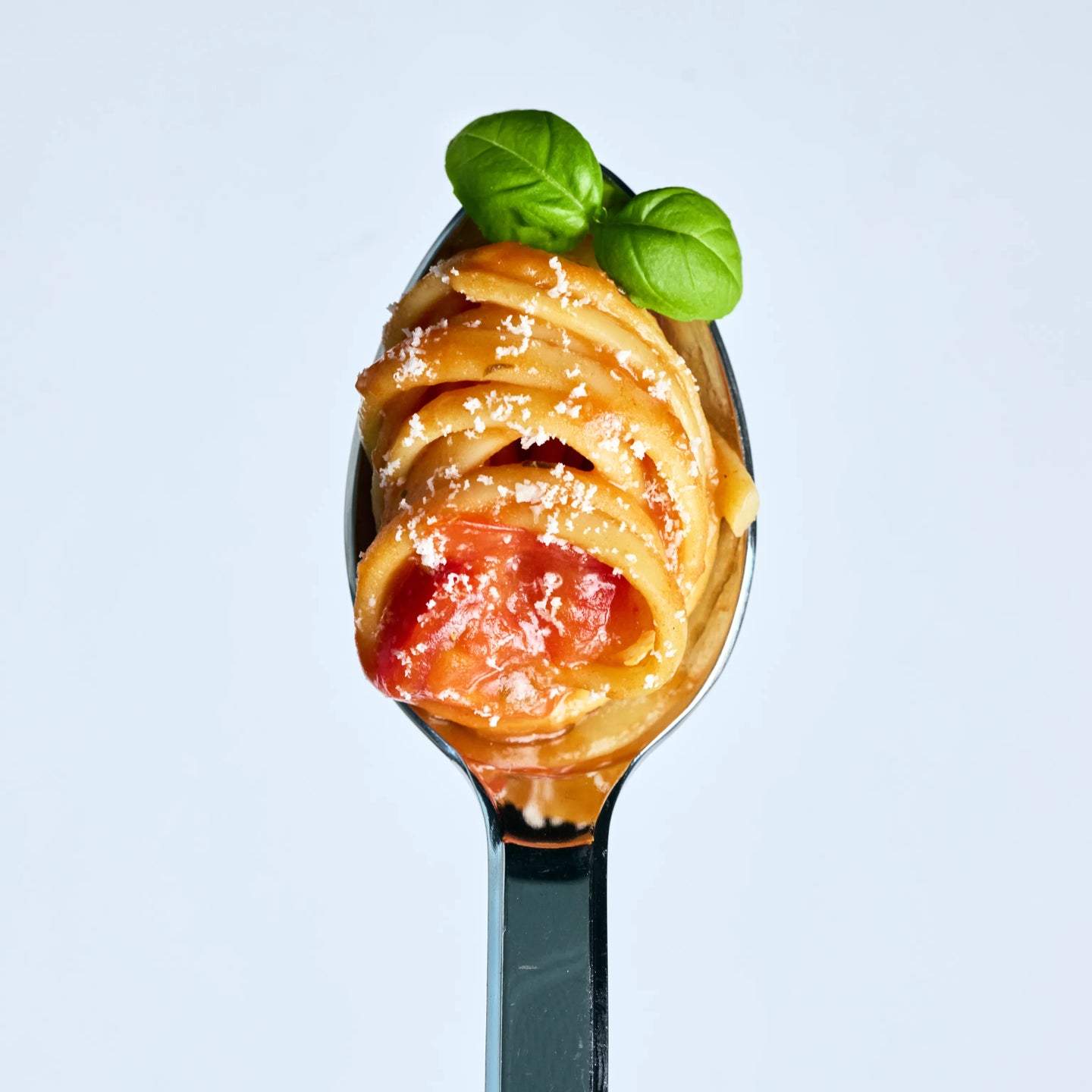 Löffel mit Spaghetti in Tomatensauce, bestreut mit Parmesan und garniert mit frischem Basilikum auf hellem Hintergrund