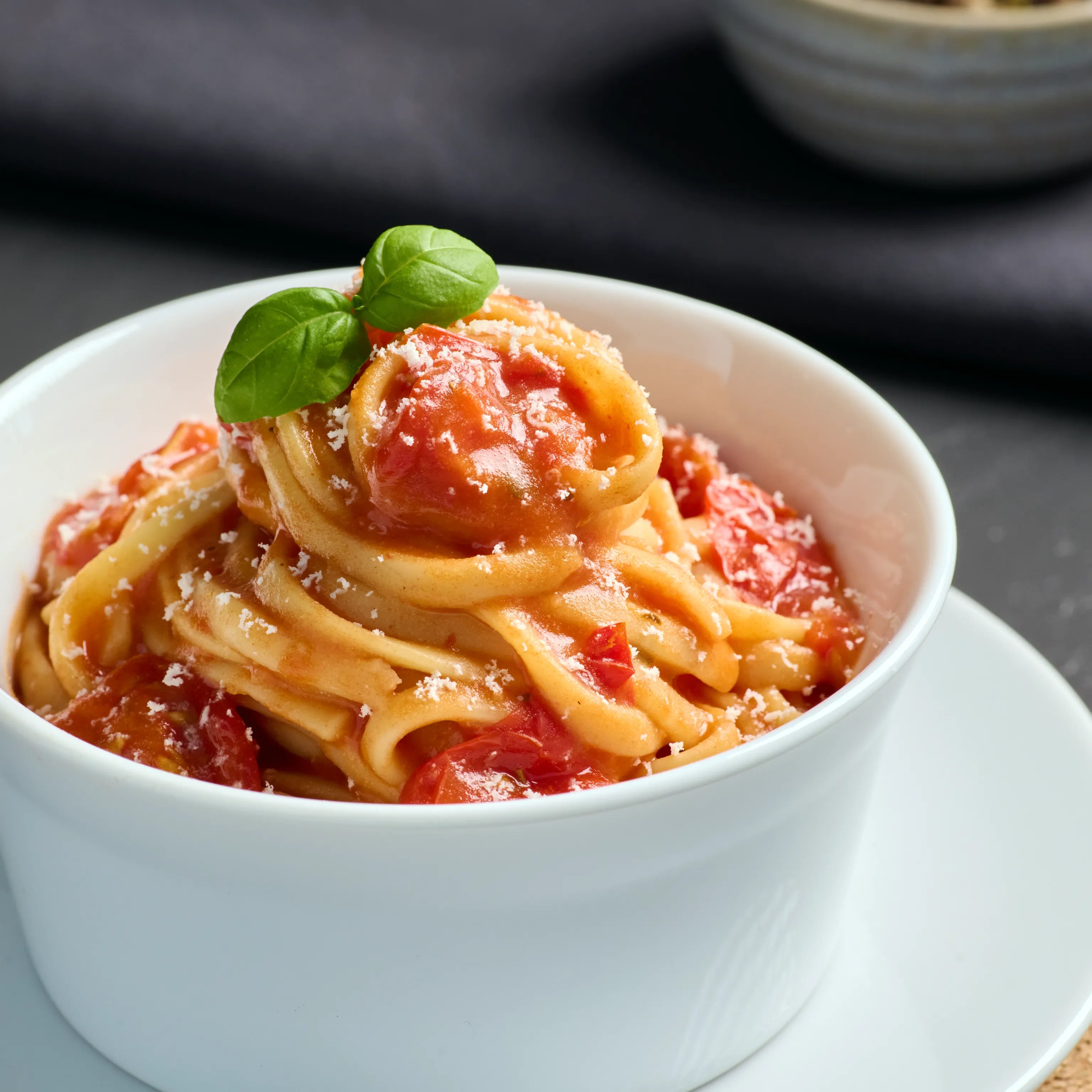 Schüssel mit Tagliatelle in Tomatensoße, garniert mit Basilikum und geriebenem Käse auf weißem Teller