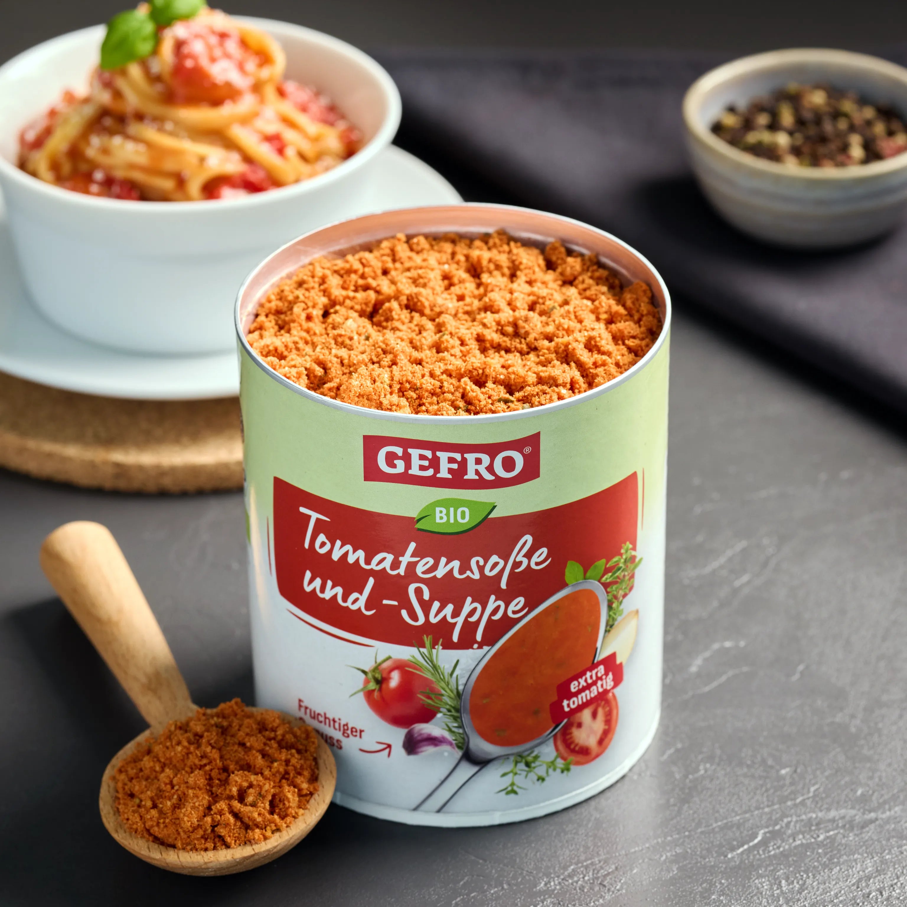 Offene Dose GEFRO Bio Tomatensoße und -Suppe mit Pulver, daneben Holzlöffel; im Hintergrund ein Teller Pasta.