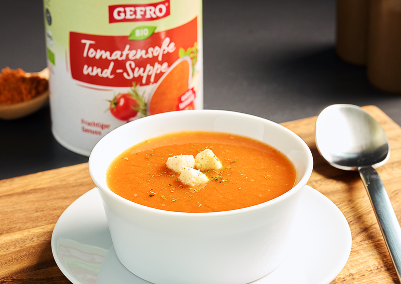 Schale mit Tomatensuppe auf Untertasse, garniert mit Croutons; dahinter Dose GEFRO Bio Tomatensoße und -Suppe
