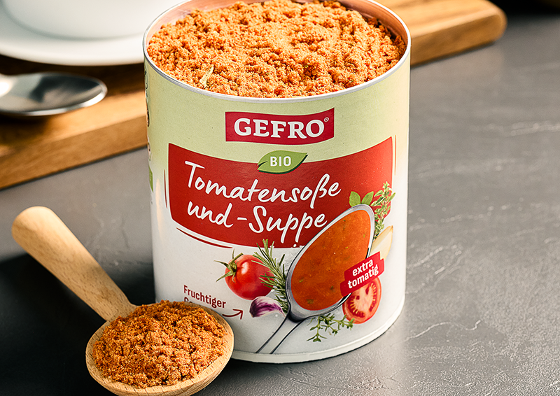 Dose GEFRO Bio Tomatensoße und -Suppe mit Pulver, daneben Holzlöffel mit Produkt zum Portionieren