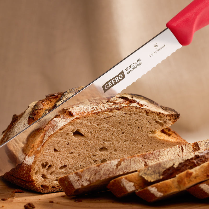 Brotmesser mit rotem Griff schneidet ein rustikales Bauernbrot auf Holzbrett, mehrere Scheiben liegen vorne bereit.