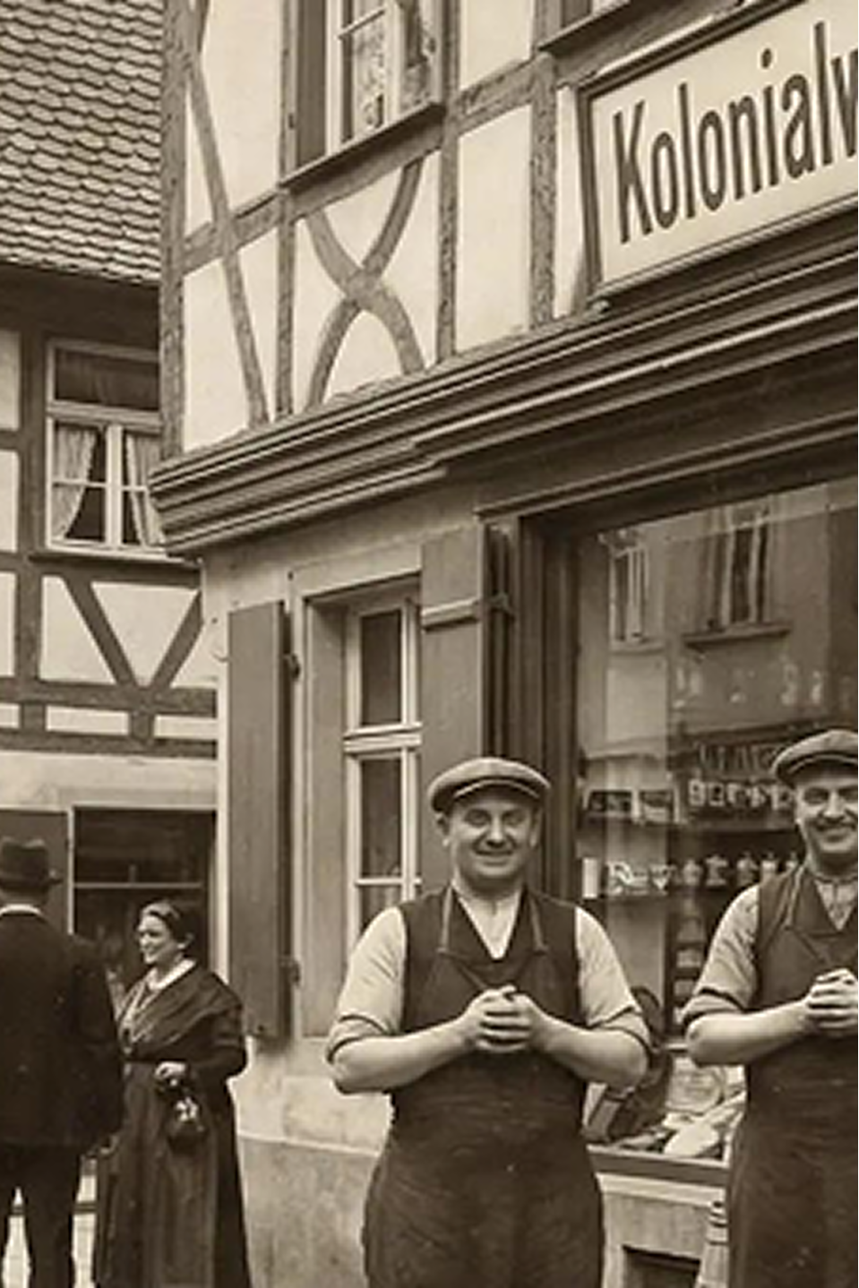 Historische Straßenszene vor einem Kolonialwarenladen mit zwei Männern in Schürzen vor Fachwerkhäusern