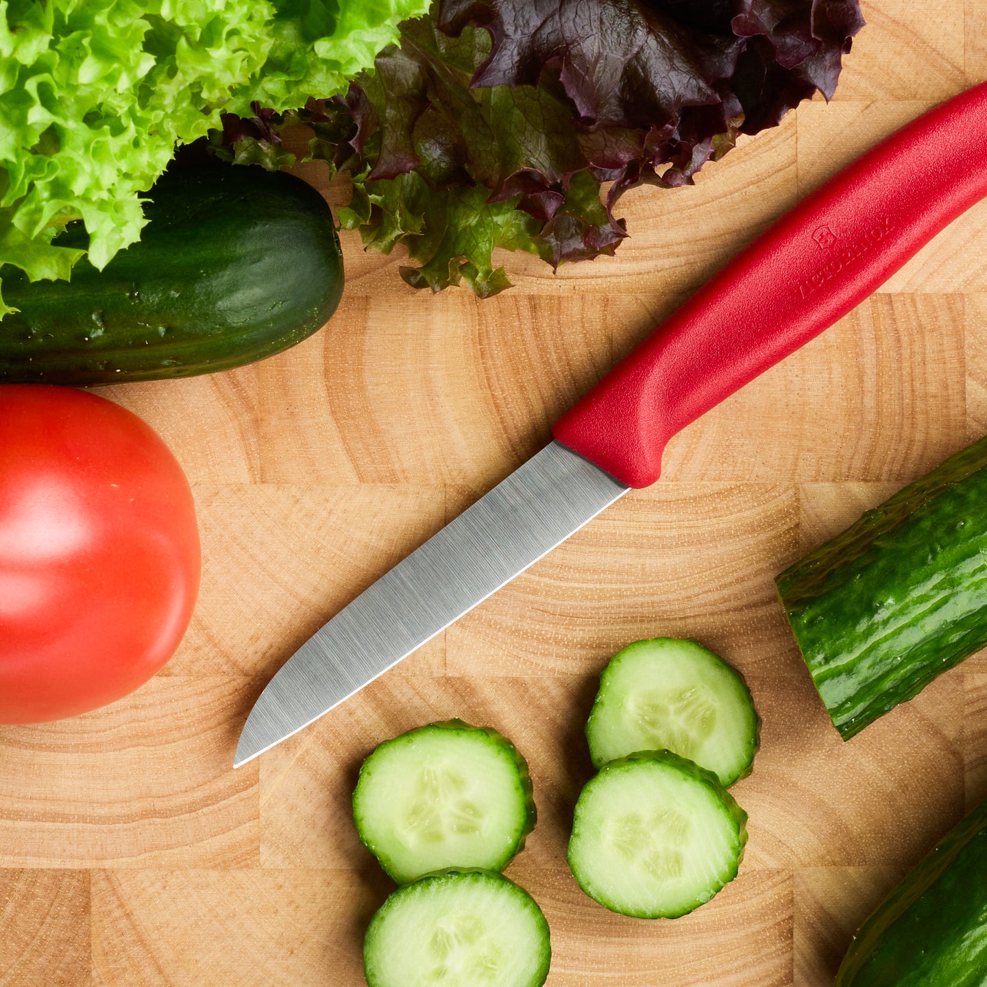 Küchenmesser mit rotem Griff auf Holzbrett, umgeben von Gurke, Tomate und Salat für die Zubereitung frischer Speisen