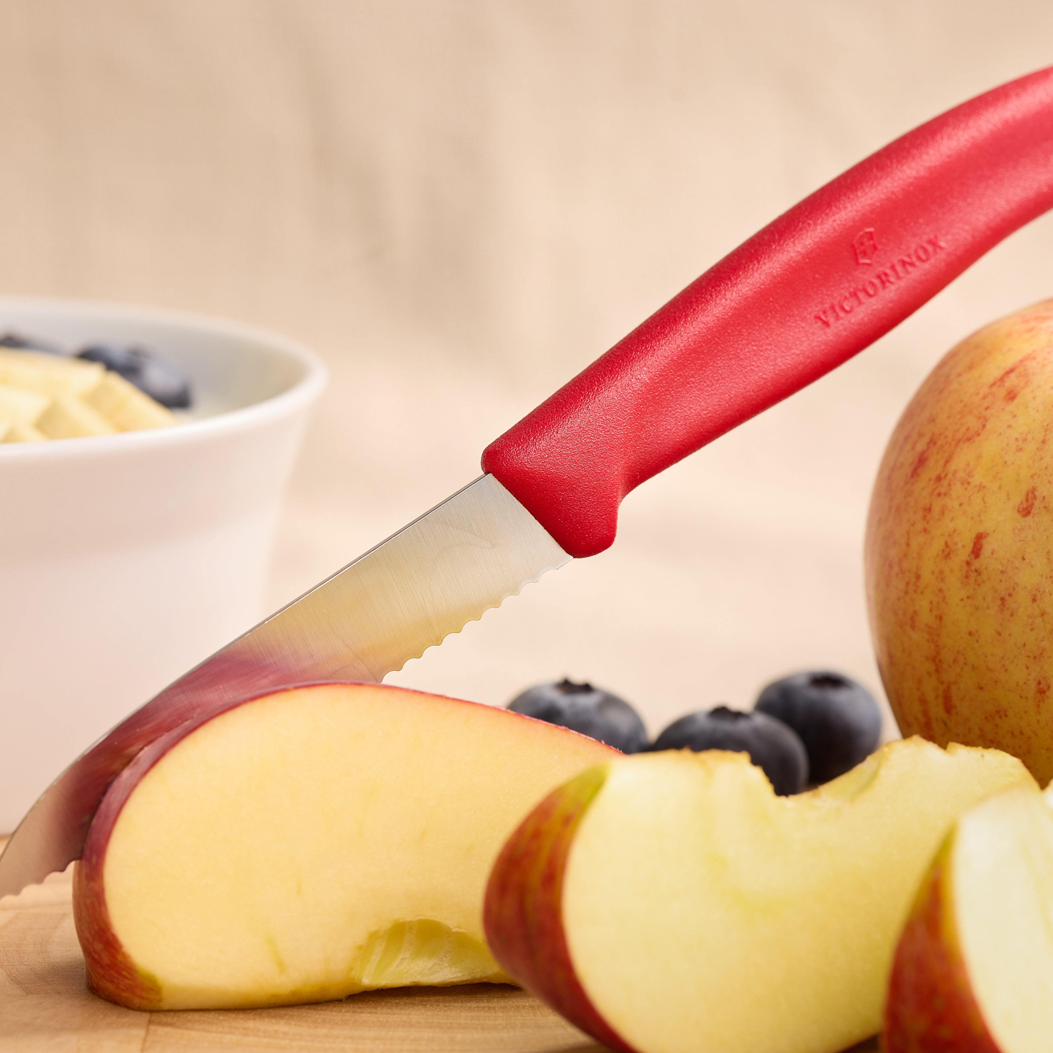 Rotes Victorinox-Obstmesser mit Wellenschliff beim Schneiden von Apfelstücken auf Holzbrett, daneben Blaubeeren