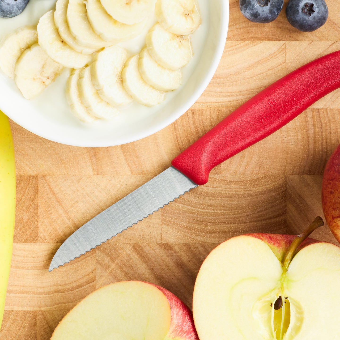Rotes Messer mit Wellenschliff auf Holzbrett neben geschnittener Banane, Äpfeln und Heidelbeeren.