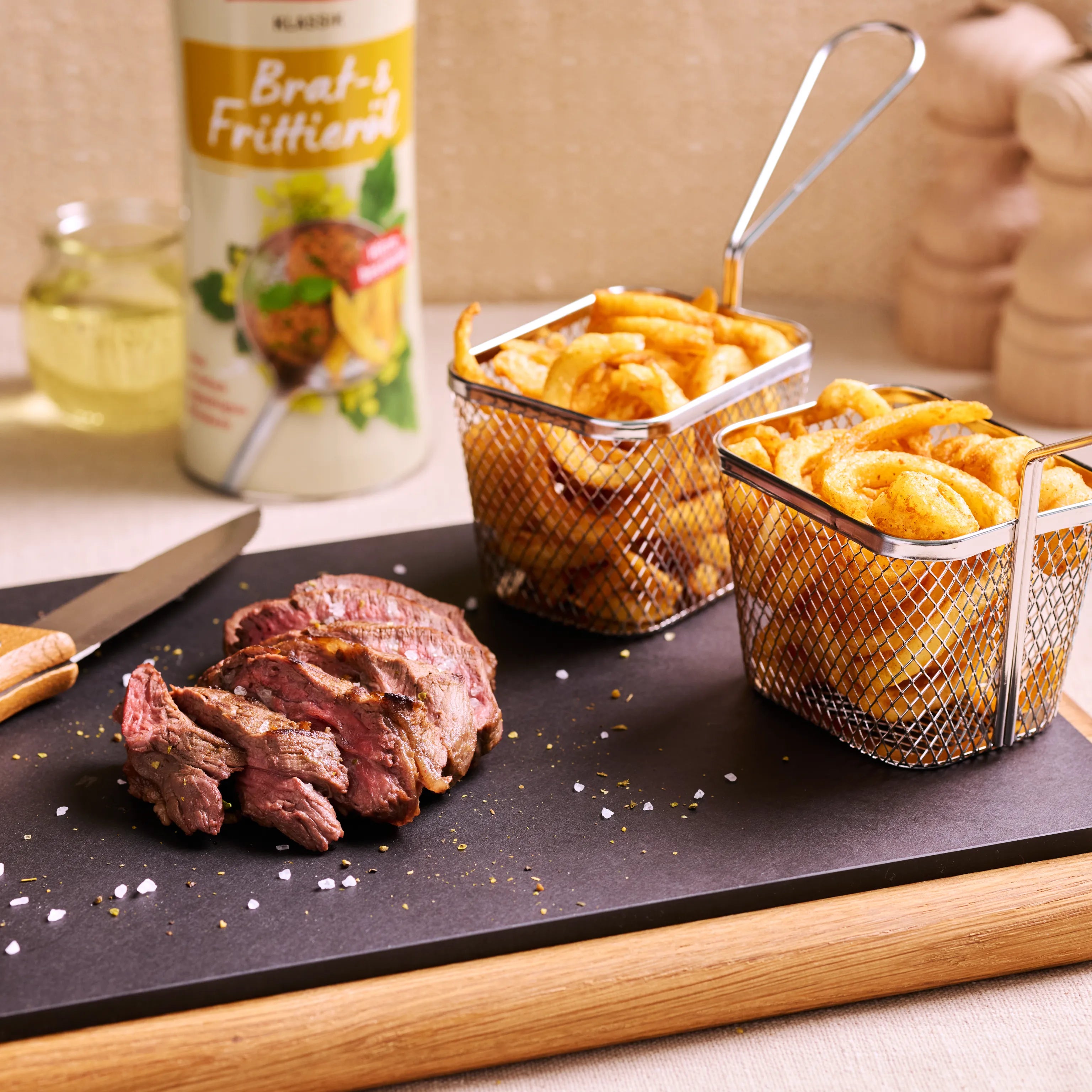Zwei Körbchen mit Pommes und Kartoffelecken neben geschnittenem Steak auf Servierbrett, im Hintergrund Bratfrittieröl