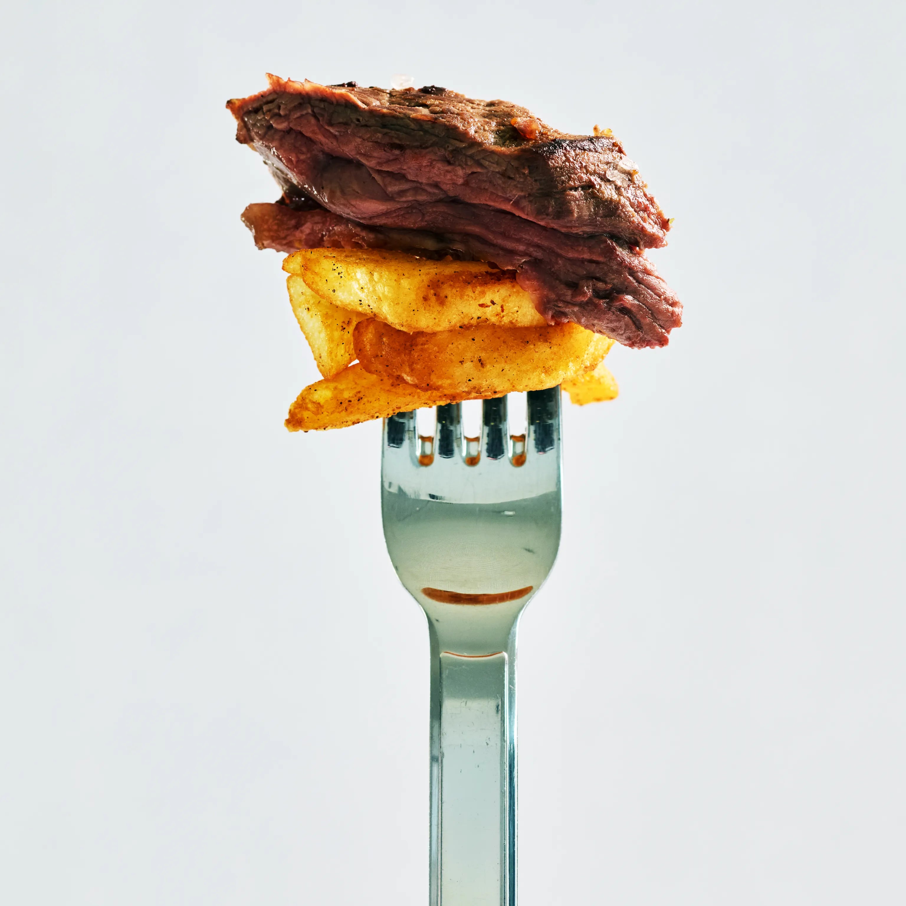 Gabel mit einem Stück gebratenem Fleisch und gewürzten Kartoffelchips vor hellem Hintergrund, Nahaufnahme