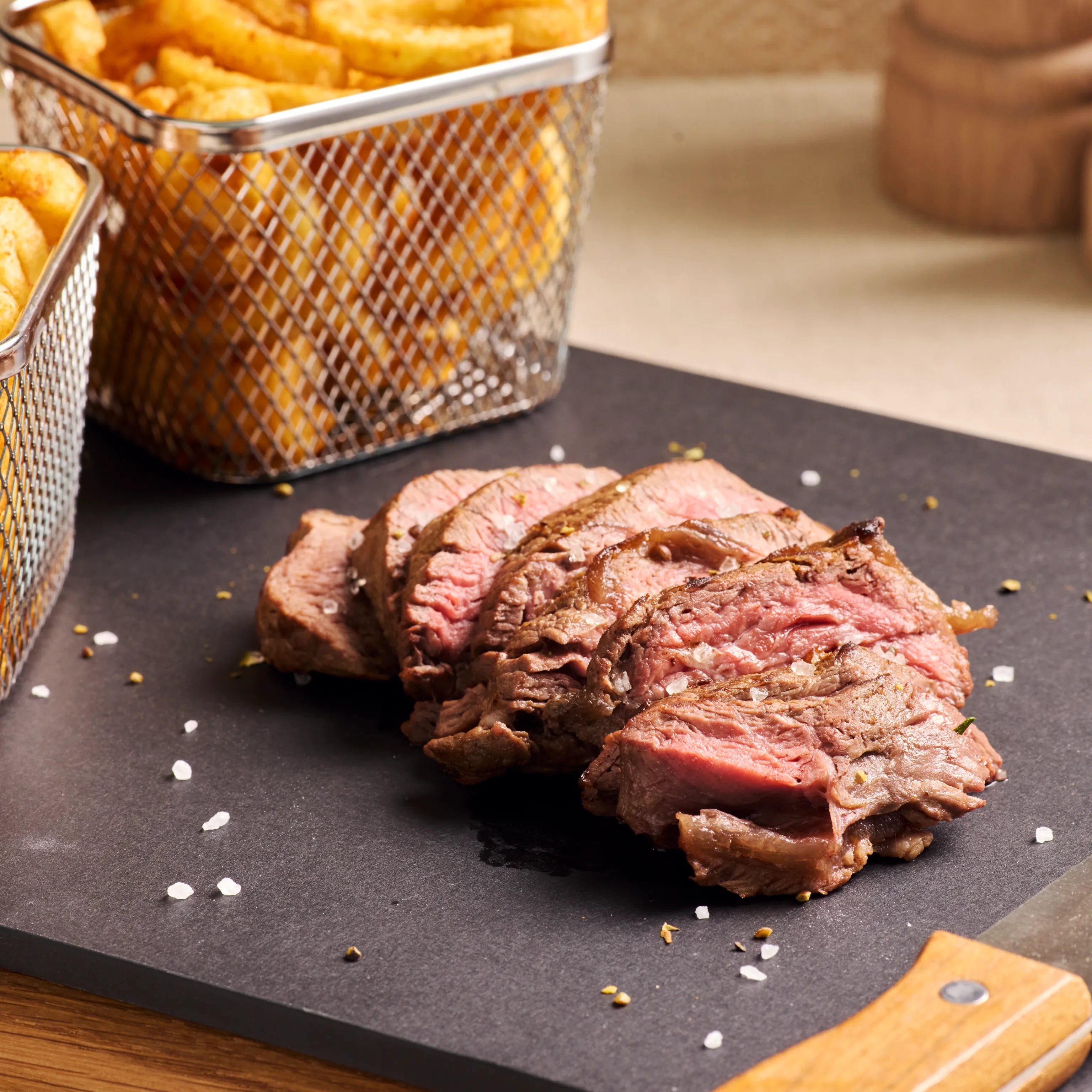 In Scheiben geschnittenes rosa Rindersteak auf schwarzer Platte, dazu Pommes in Metallkörben im Hintergrund
