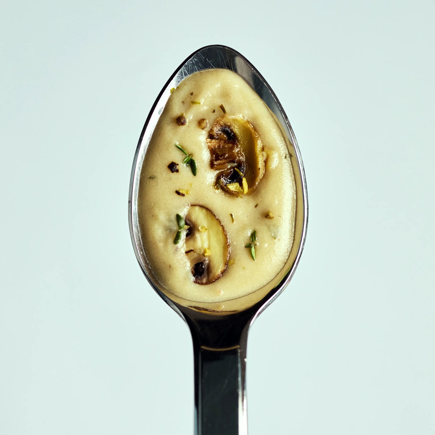 Löffel mit cremiger Pilzsuppe, garniert mit Champignonscheiben und Kräutern, vor hellem Hintergrund dargestellt