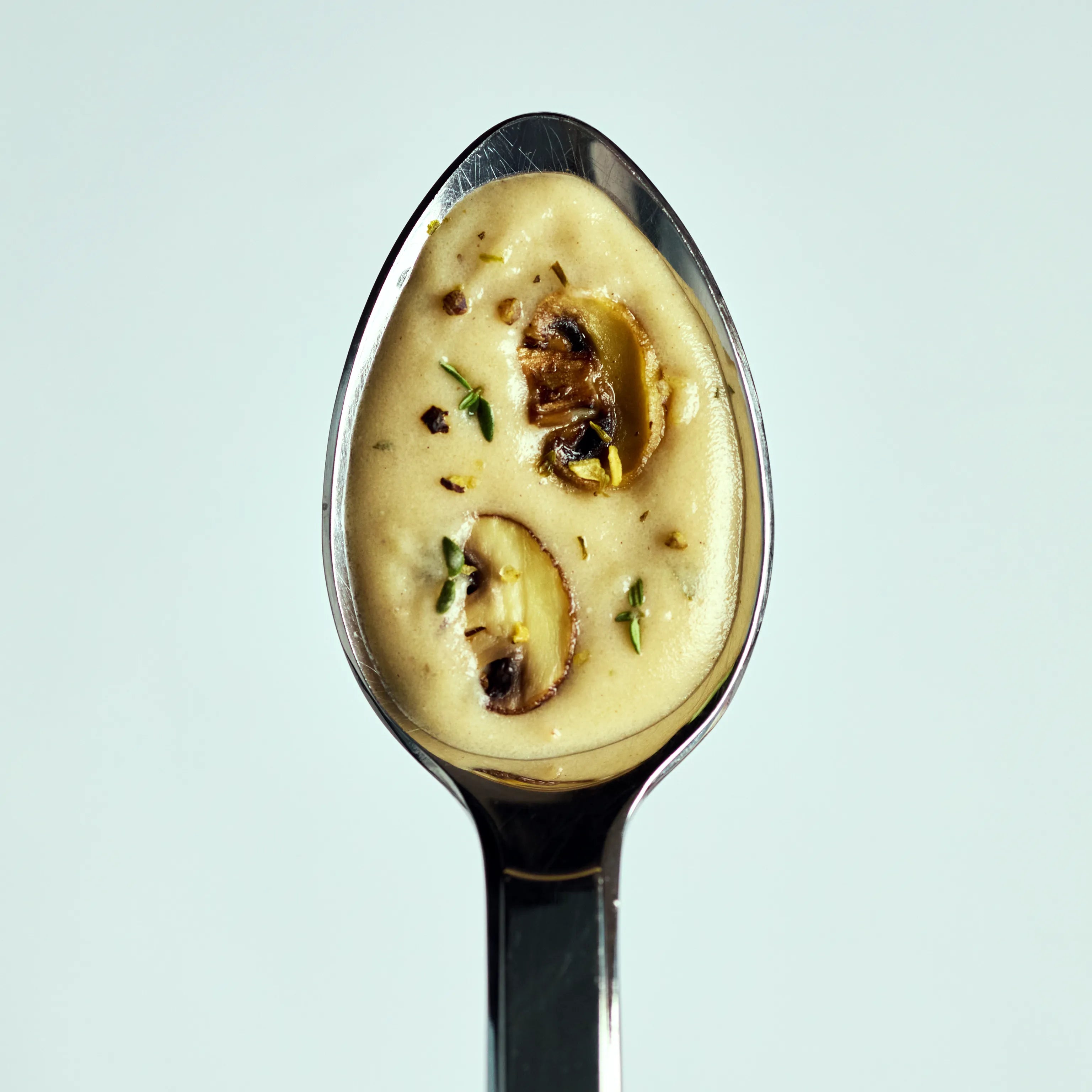 Löffel mit cremiger Pilzsuppe, garniert mit Champignonscheiben und Kräutern, vor hellem Hintergrund dargestellt