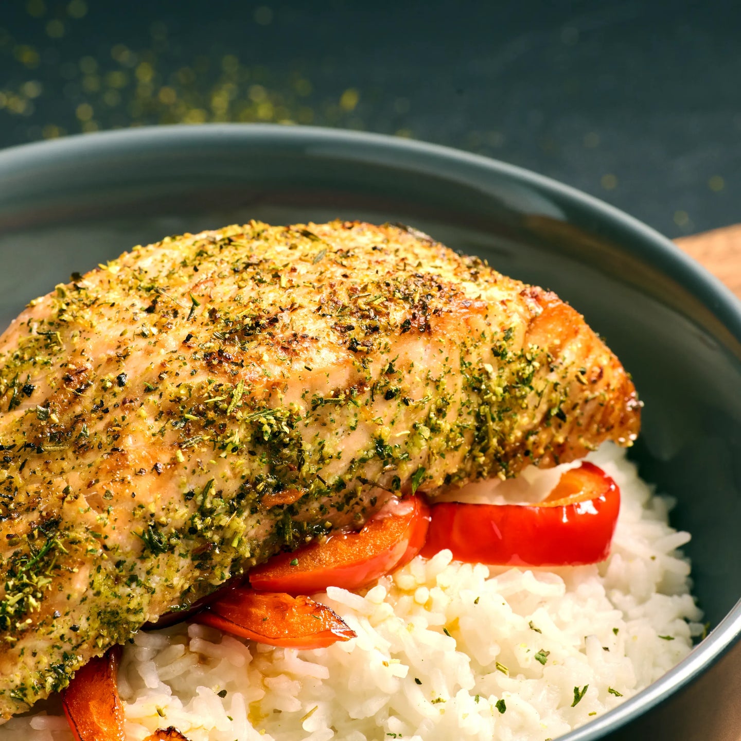 Gebratenes Hähnchenfilet mit Kräutern auf weißem Reis und roter Paprika in einer dunklen Schale serviert