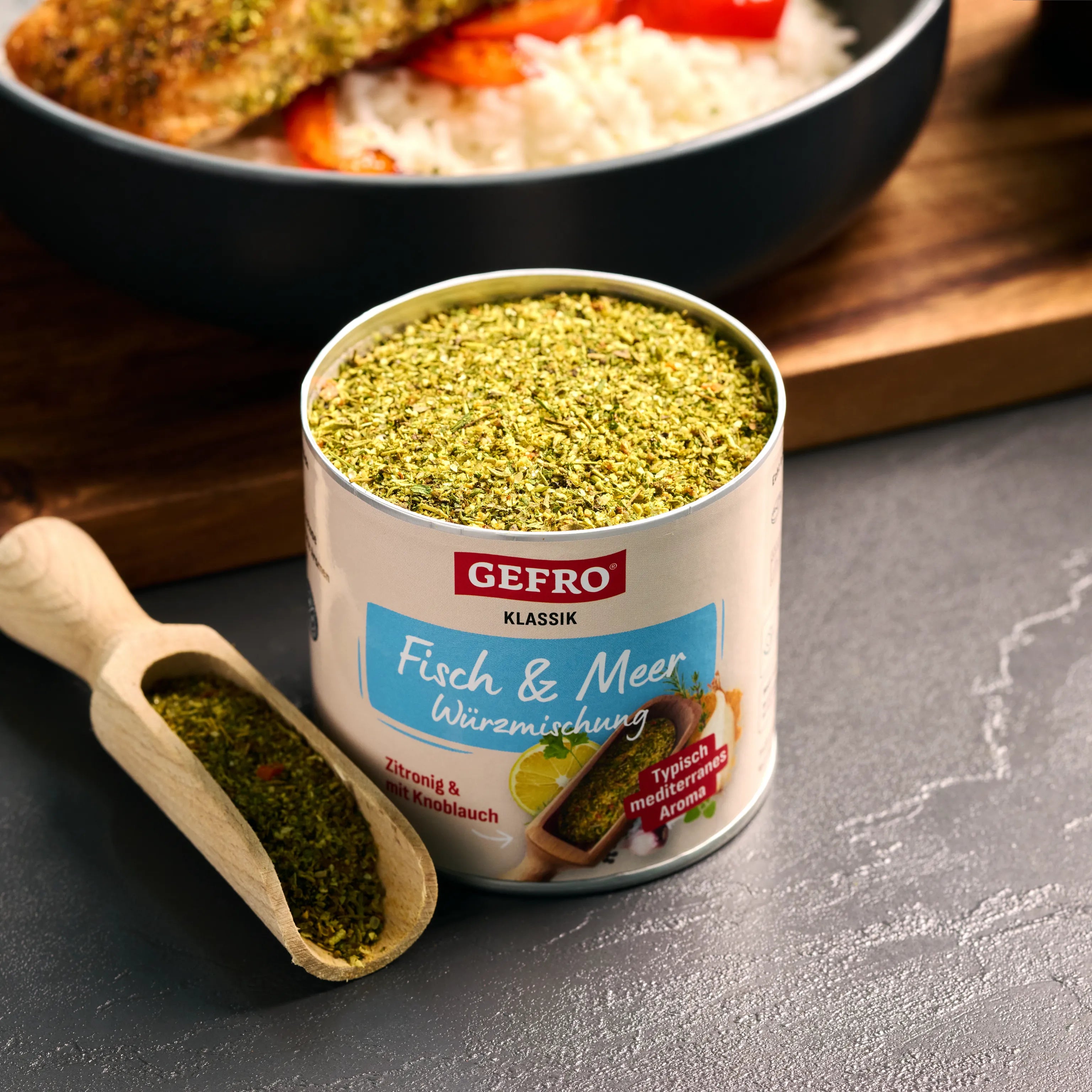 Offene Dose GEFRO Fisch & Meer Würzmischung mit Holzschaufel; im Hintergrund ein Fischgericht mit Reis und Gemüse.