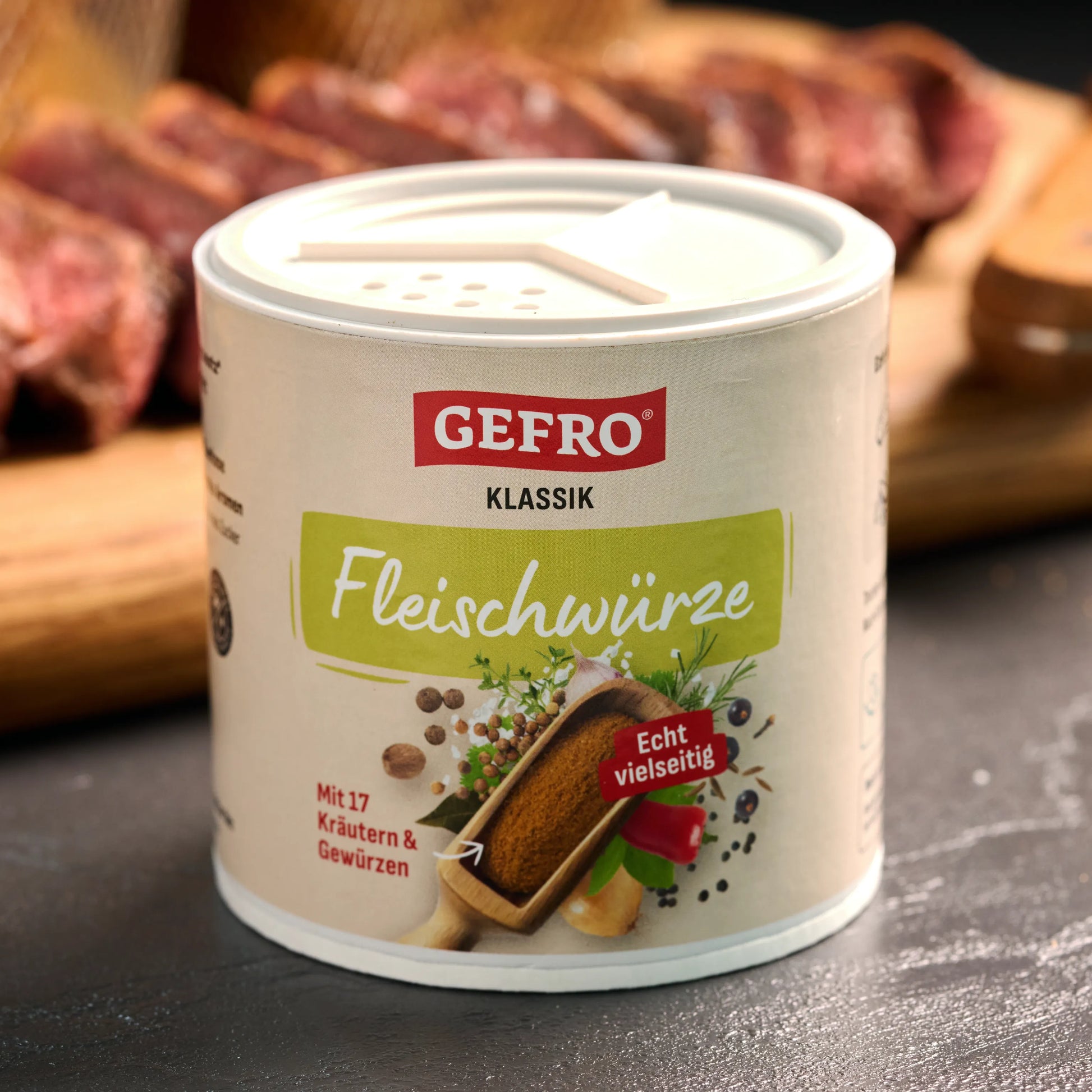 Dose GEFRO Klassik Fleischwürze mit Streudeckel, davor aufgestellt, im Hintergrund rohes Fleisch auf Holzbrett.