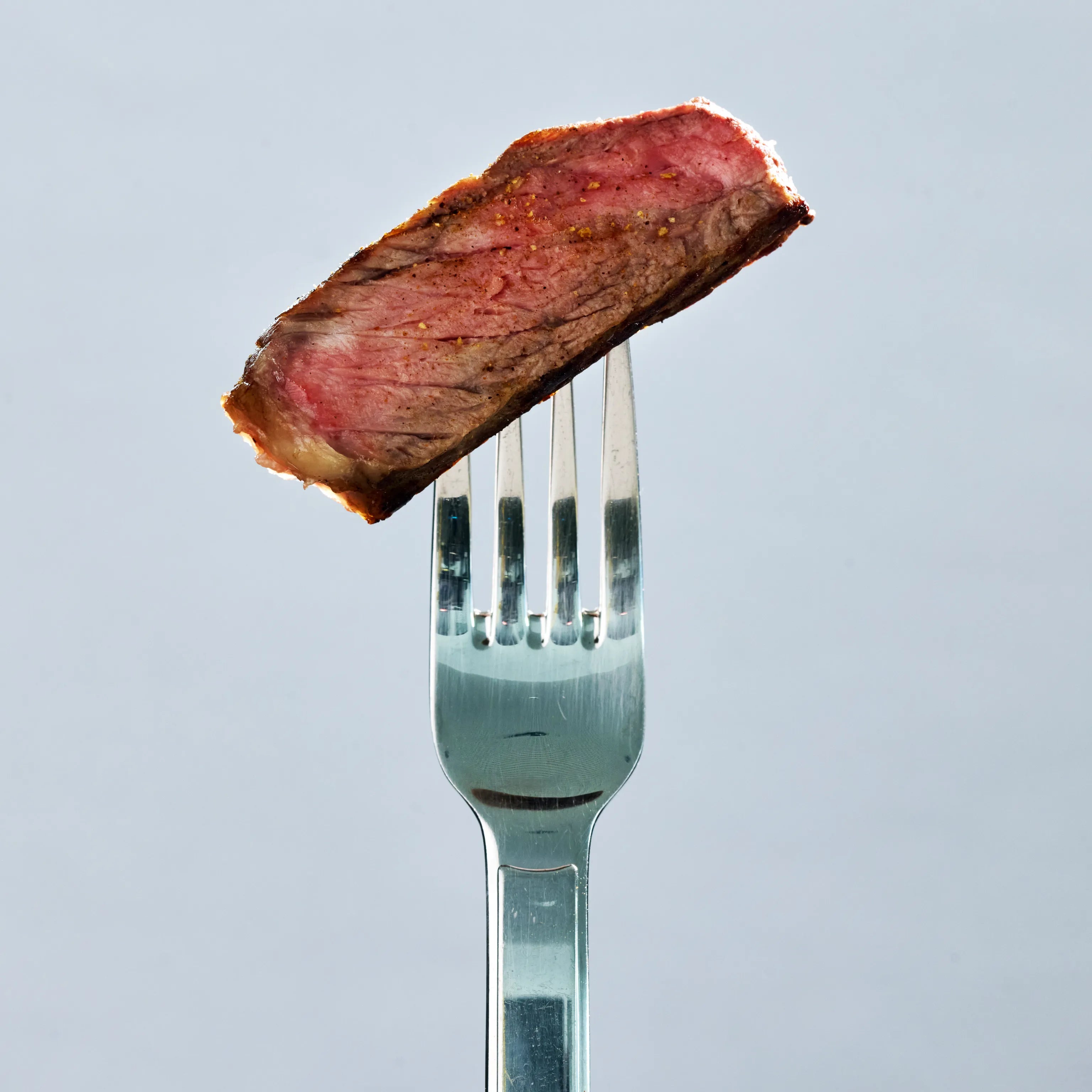 Nahaufnahme eines rosa gebratenen Steakstücks auf einer Gabel, saftig mit gebräunter Kruste vor hellem Hintergrund