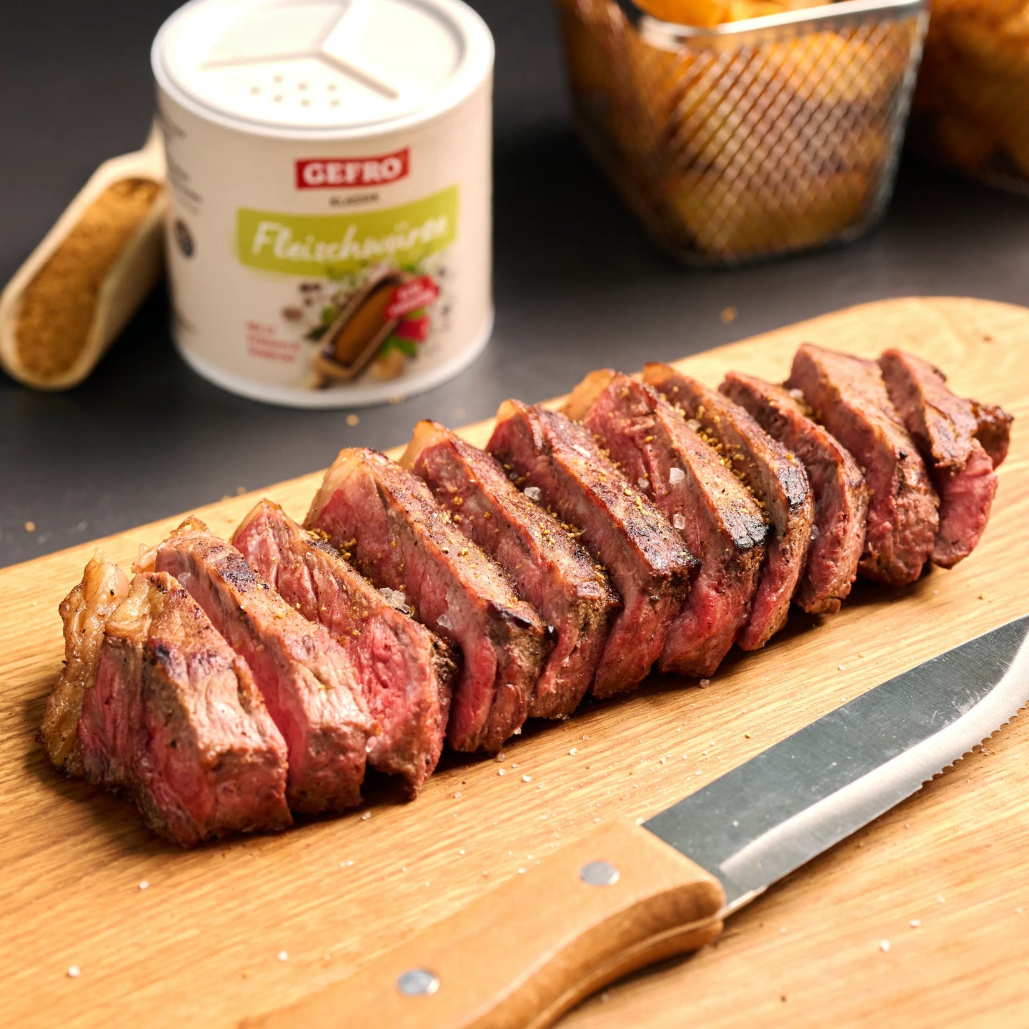 In Scheiben geschnittenes, rosa gebratenes Steak auf Holzbrett, daneben Messer und Dose Fleischgewürz.
