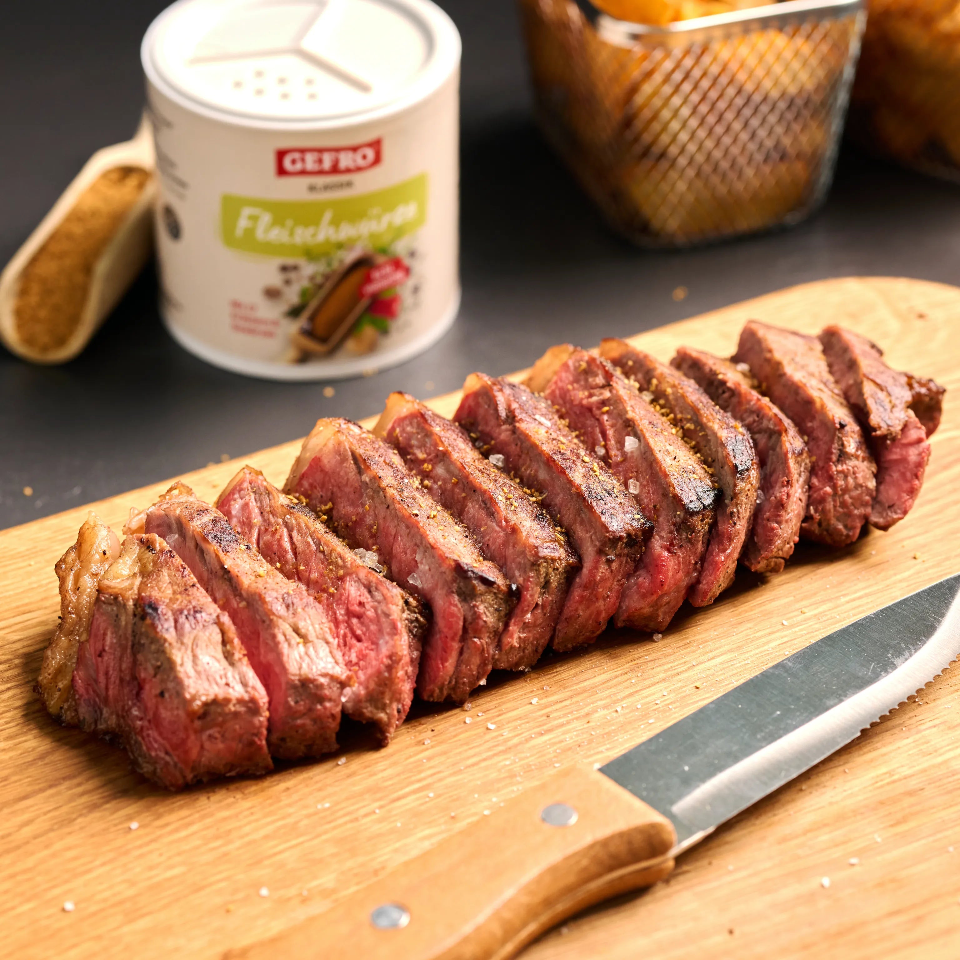 In Scheiben geschnittenes, rosa gebratenes Steak auf Holzbrett, daneben Messer und Dose Fleischgewürz.