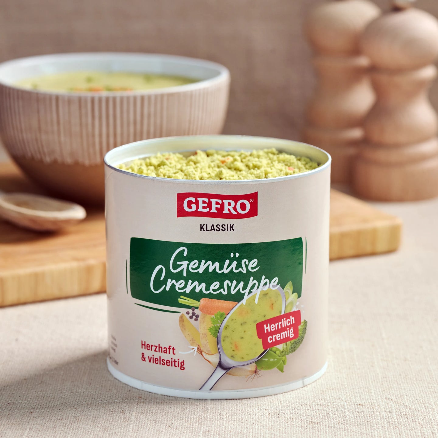 Dose GEFRO Klassik Gemüse-Cremesuppe mit Pulver, davor auf Tisch, im Hintergrund eine Schüssel mit fertiger Suppe.
