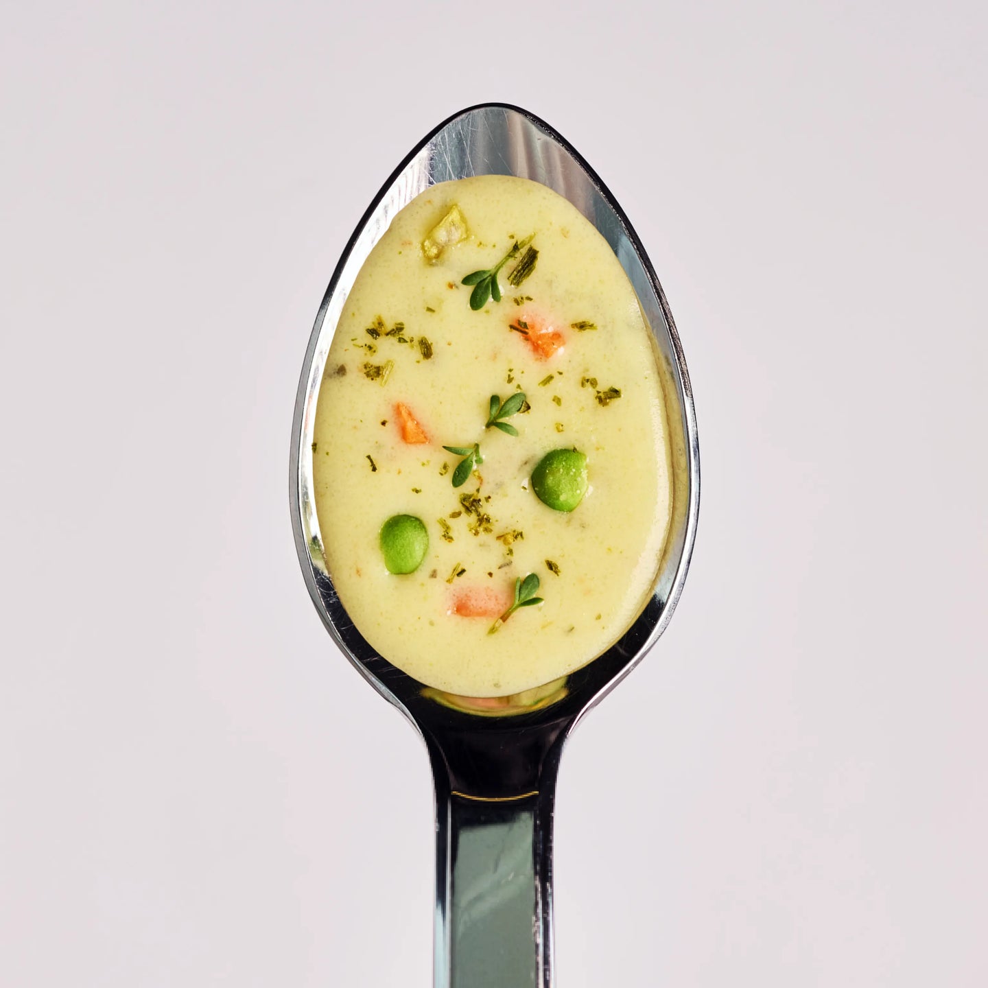 Löffel mit cremiger Gemüsesuppe, garniert mit Erbsen, Kräutern und kleinen Gemüsestücken auf hellem Hintergrund