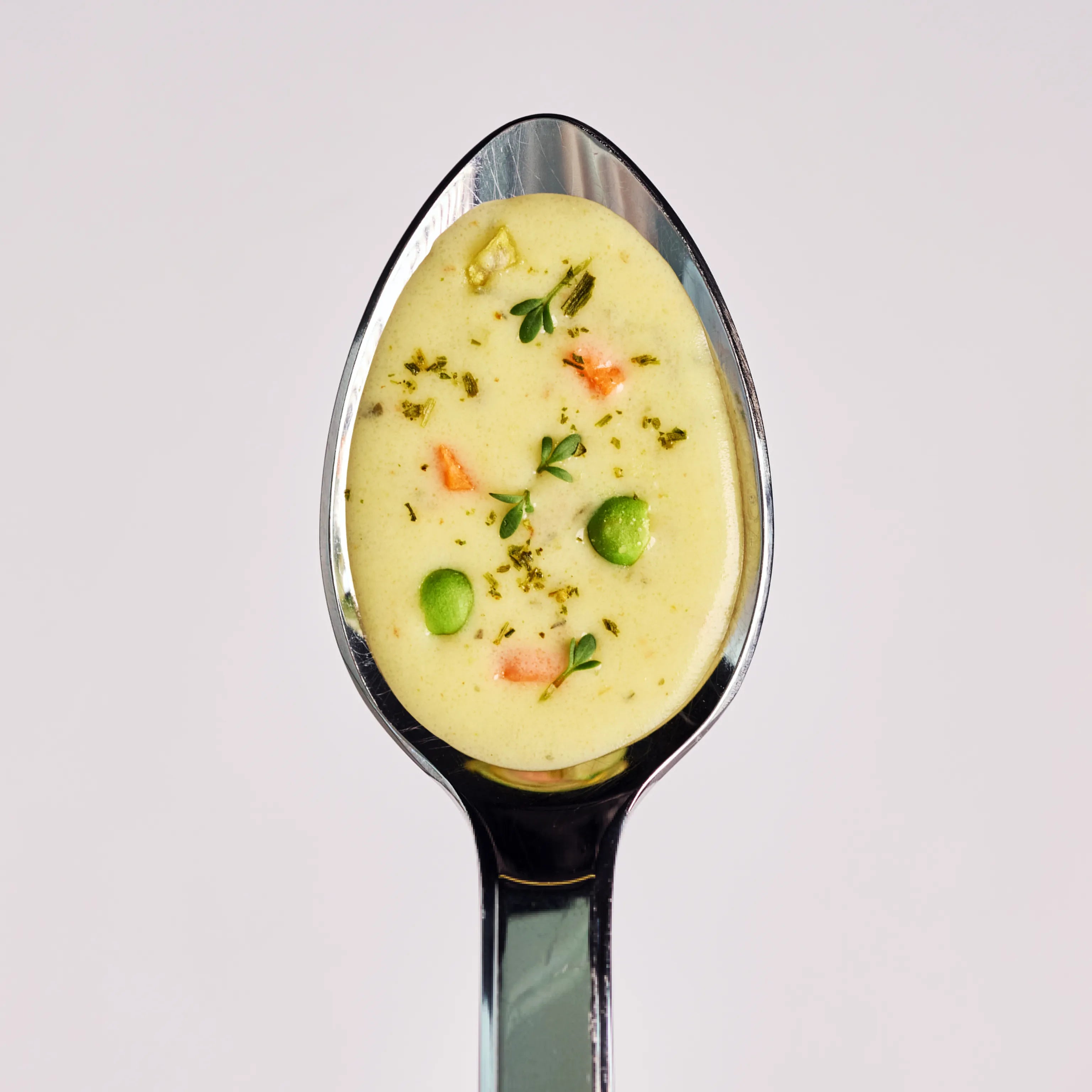 Löffel mit cremiger Gemüsesuppe, garniert mit Erbsen, Kräutern und kleinen Gemüsestücken auf hellem Hintergrund